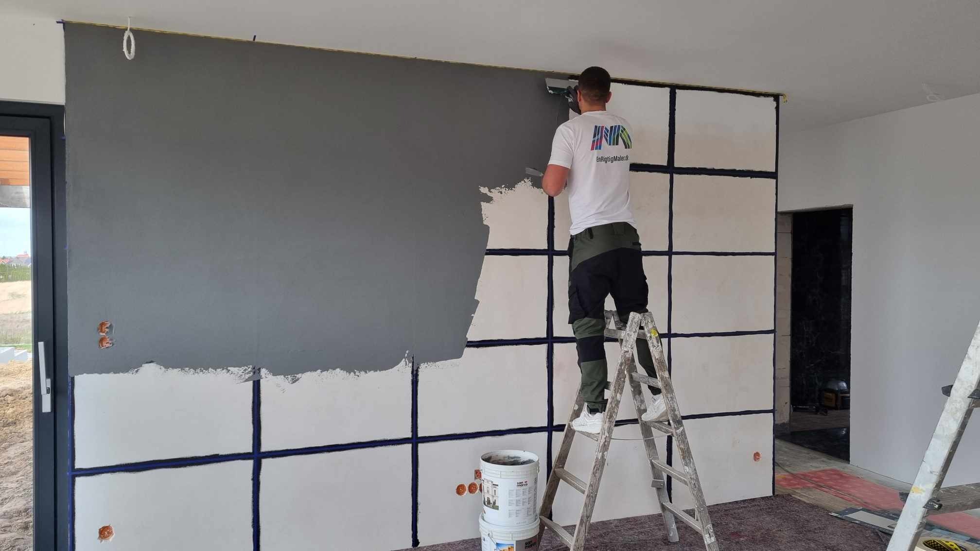 Pracownik na drabinie aplikuje szarą farbę na ścianę w pomieszczeniu, widoczne puszki z farbą i częściowo pomalowana ściana z zaznaczonymi liniami