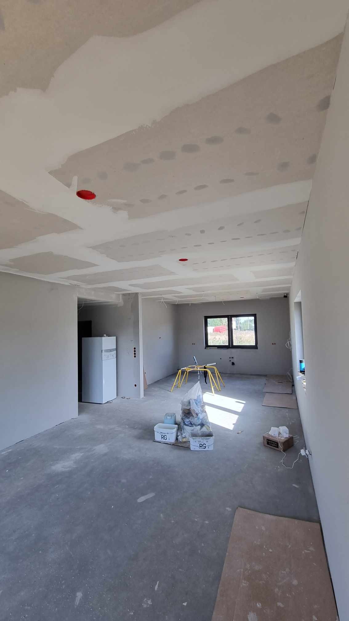 Wnętrze w trakcie prac wykończeniowych: sufit z widocznymi fragmentami płyt gipsowo-kartonowych przed ostatecznym wykończeniem, ściany w szarym kolorze, na podłodze resztki materiałów budowlanych...