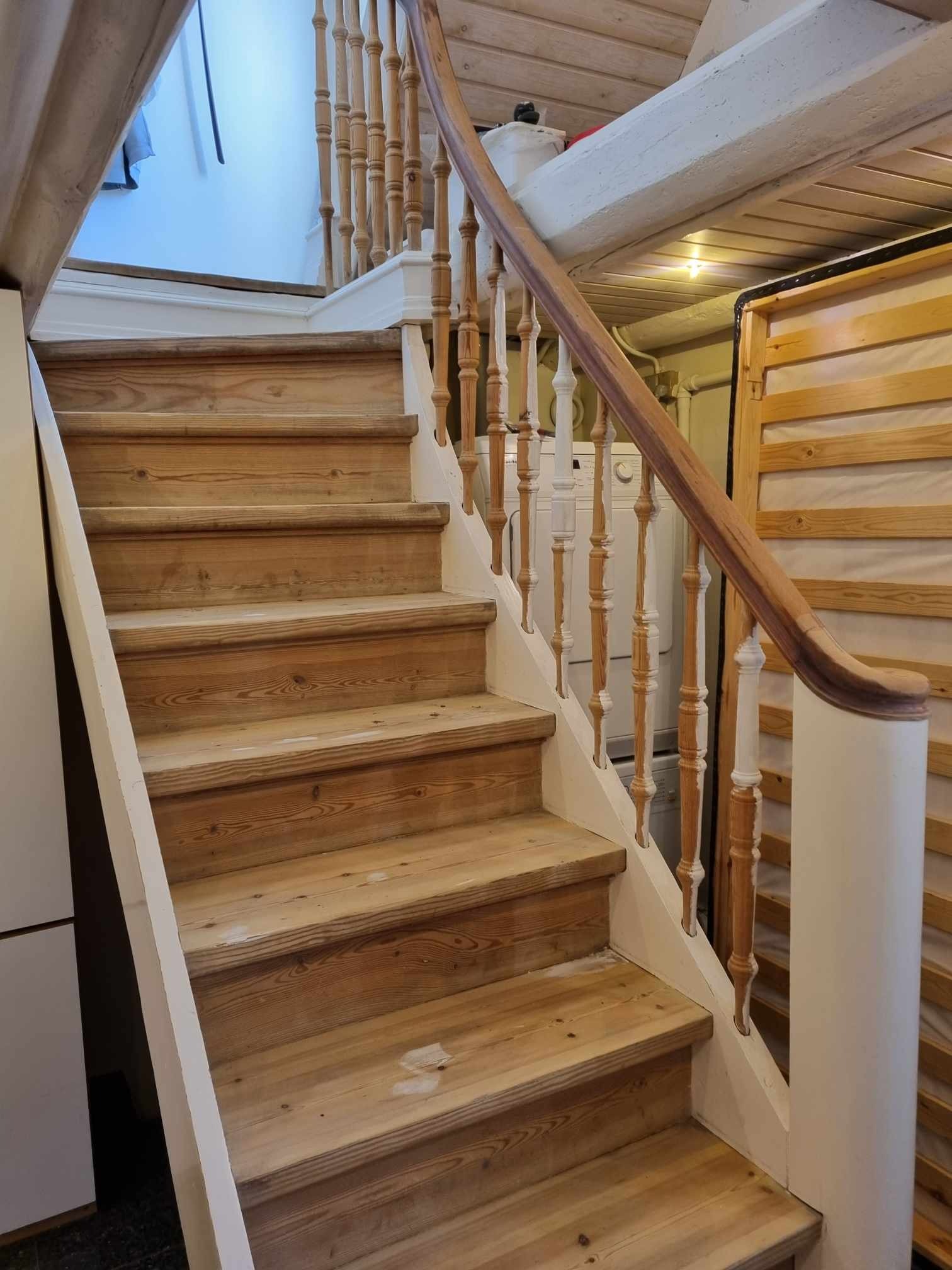 Drewniane schody z balustradą w trakcie renowacji, widoczne ślady po usuniętej farbie i niedomalowane elementy, w tle pralka i płyta meblowa.