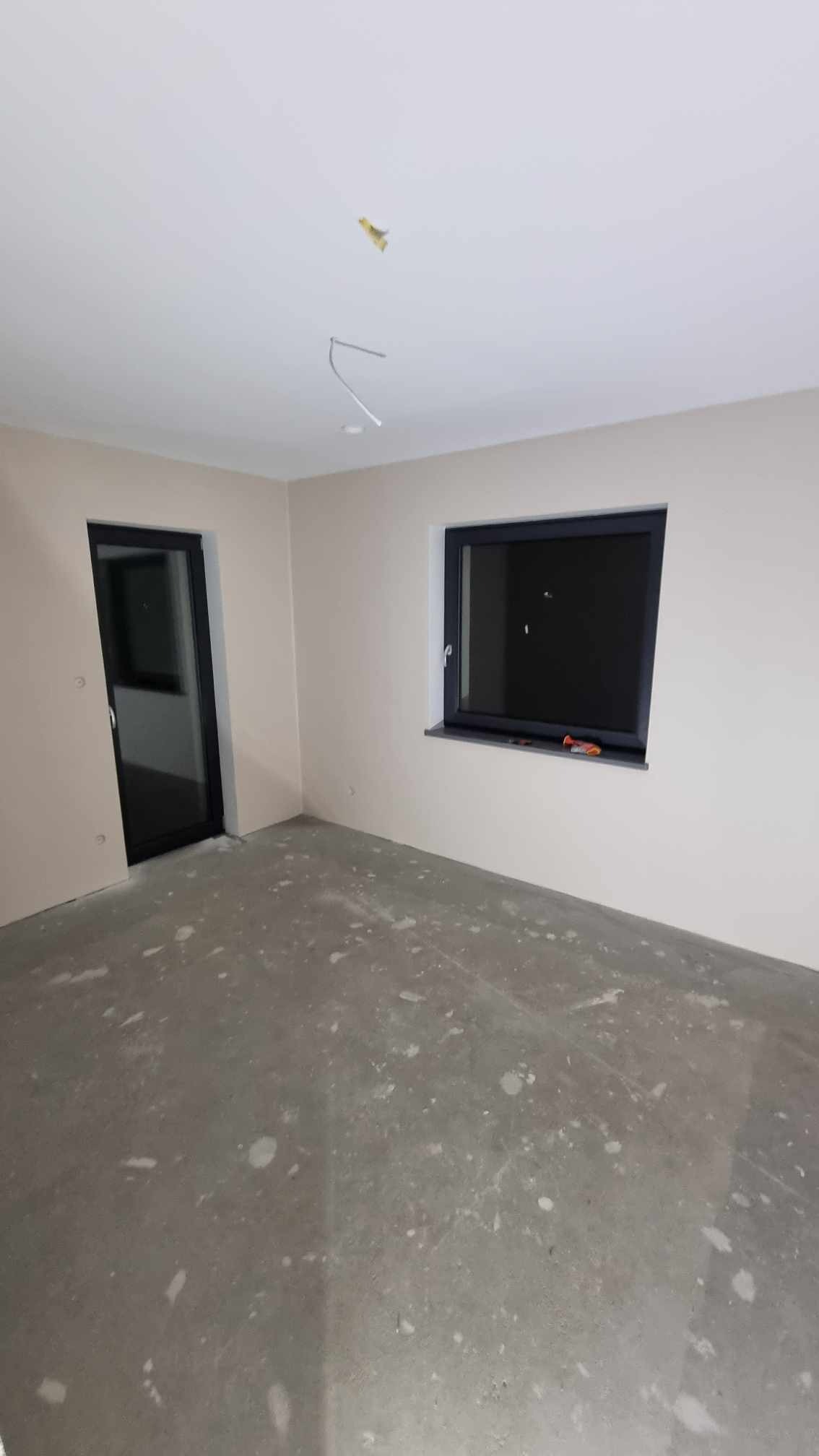 Pokój w trakcie prac wykończeniowych, ściany pomalowane na jasny kolor, surowa wylewka na podłodze, widoczne przewody elektryczne wystające z sufitu, okno z ciemną ramą.