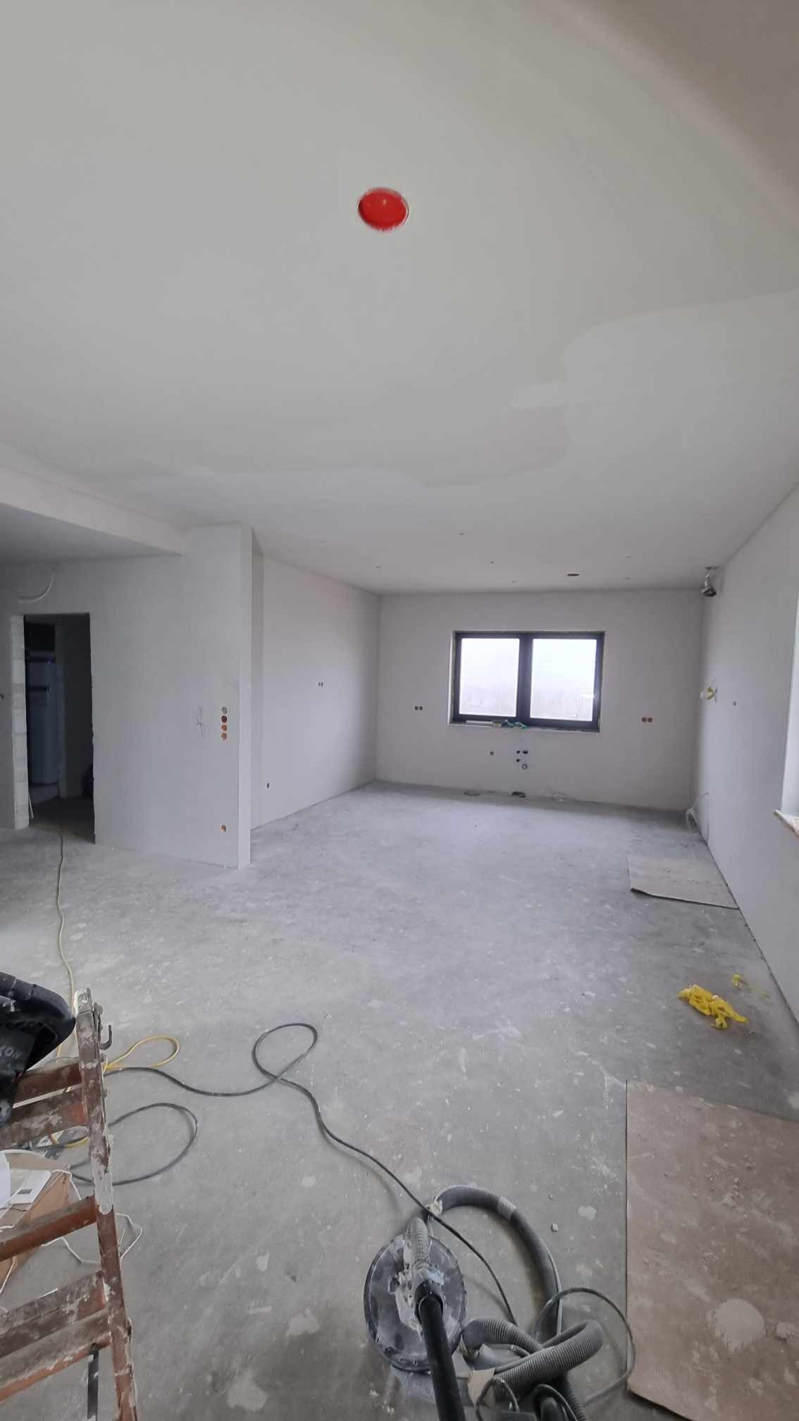 Wnętrze w trakcie prac wykończeniowych: szlifowanie podłogi i ścian, widoczny szlifierka, drabina malarska i resztki materiałów budowlanych w dużym, jasnym pomieszczeniu z oknem.