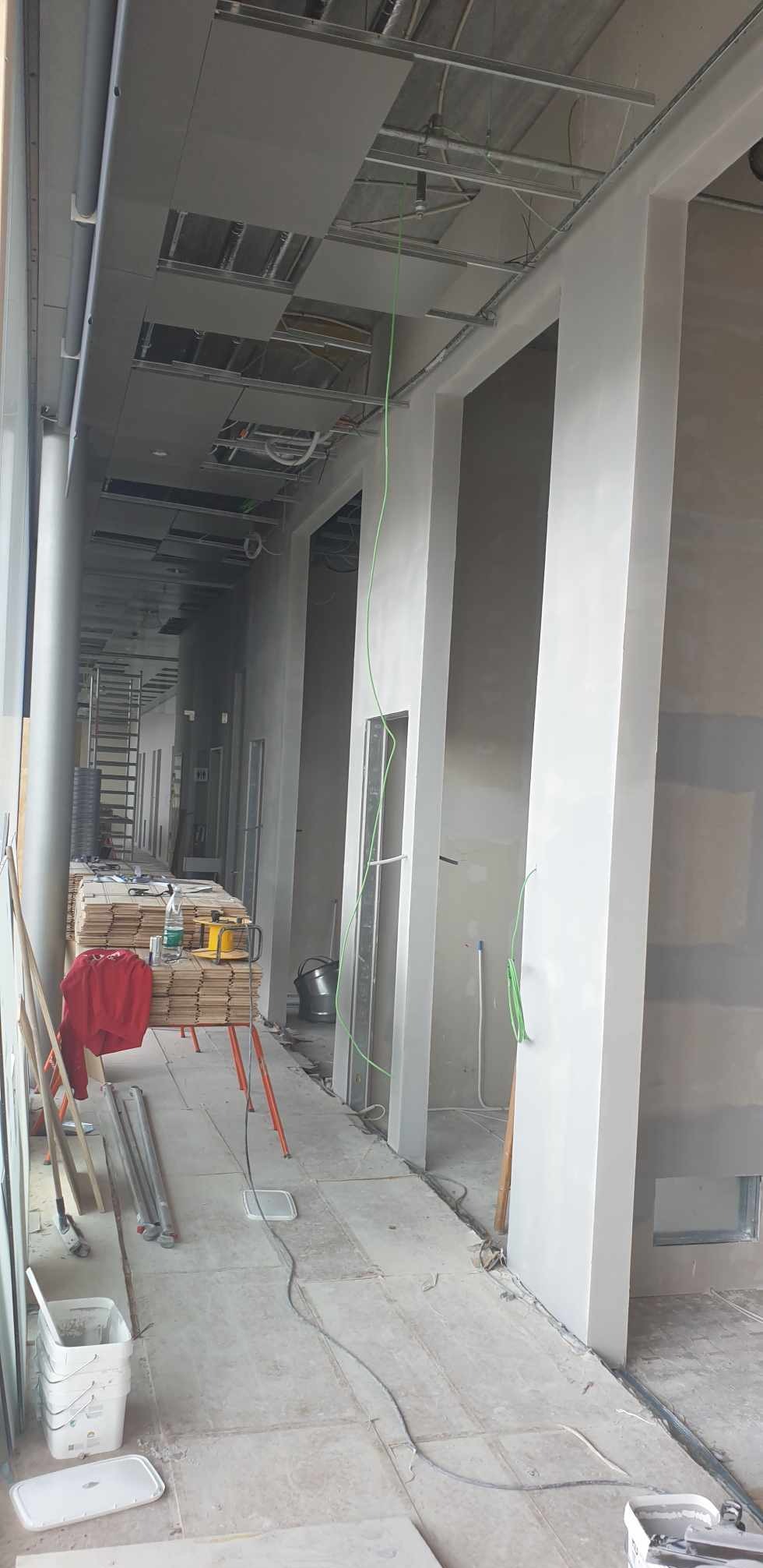 Wnętrze biurowe w trakcie prac: widoczne surowe ściany z nałożoną warstwą szpachli, odsłonięte elementy instalacji pod sufitem, materiały budowlane i narzędzia na podłodze, przewody elektryczne.