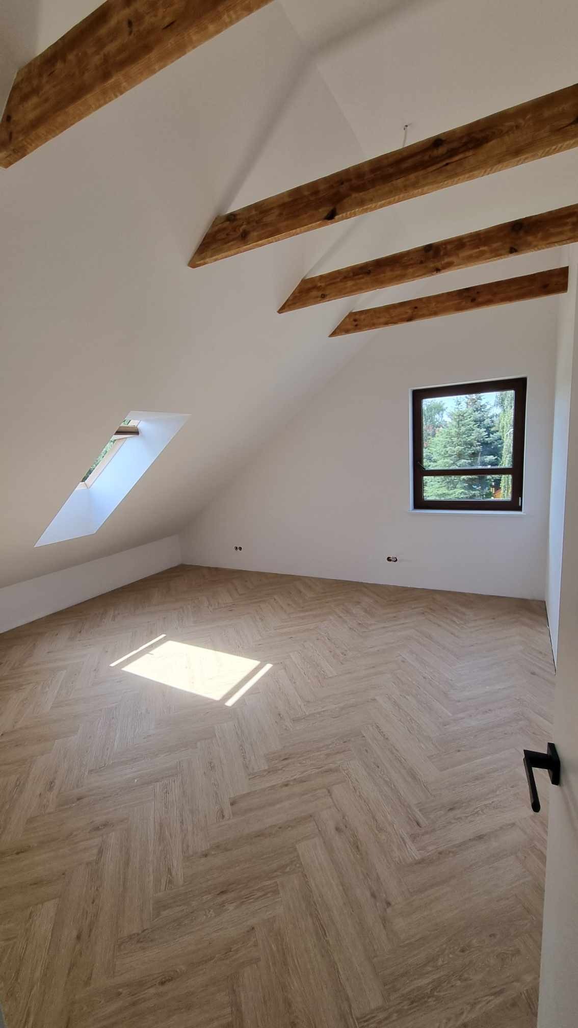 Poddasze po remoncie z drewnianymi belkami stropowymi, jasną podłogą ułożoną we wzór jodełki i oknami dachowymi oraz widokiem na zieleń.