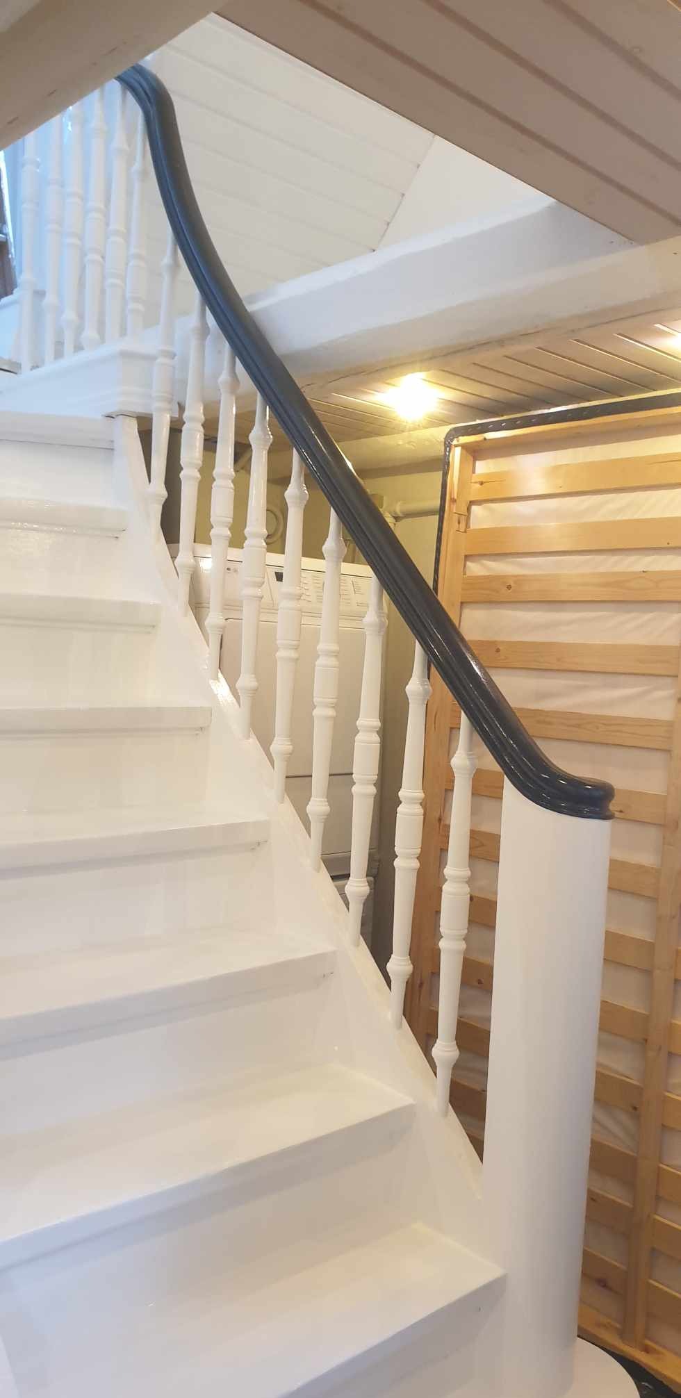 Białe schody z czarną poręczą, widok z dołu, po malowaniu, w tle pralka i drewniany stelaż łóżka.