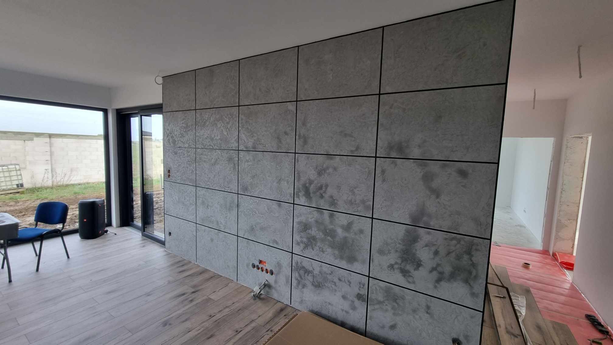 Ściana wykończona panelami dekoracyjnymi imitującymi beton, z czarnymi ramkami, widoczne gniazdka elektryczne i fragment drewnianej podłogi w surowym wnętrzu.