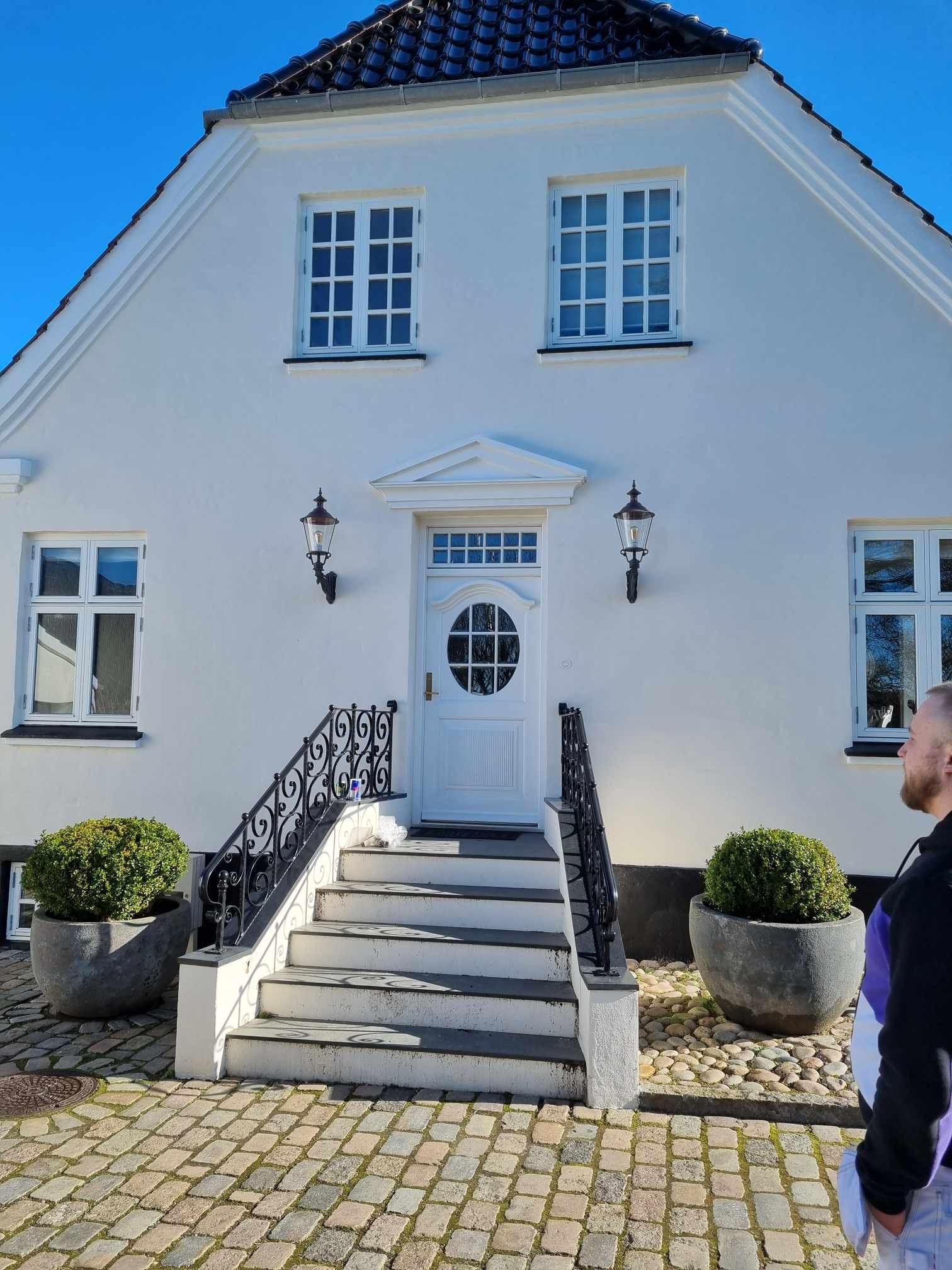 Elewacja domu z białymi ścianami, schodami z czarną balustradą i dwoma krzewami w donicach po bokach wejścia.