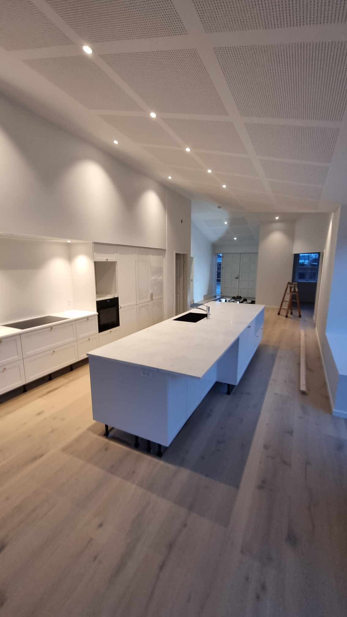 Jasna, przestronna kuchnia z wyspą i drewnianą podłogą, widoczne białe ściany, sufit z panelami akustycznymi i oświetleniem punktowym, drabina w tle.