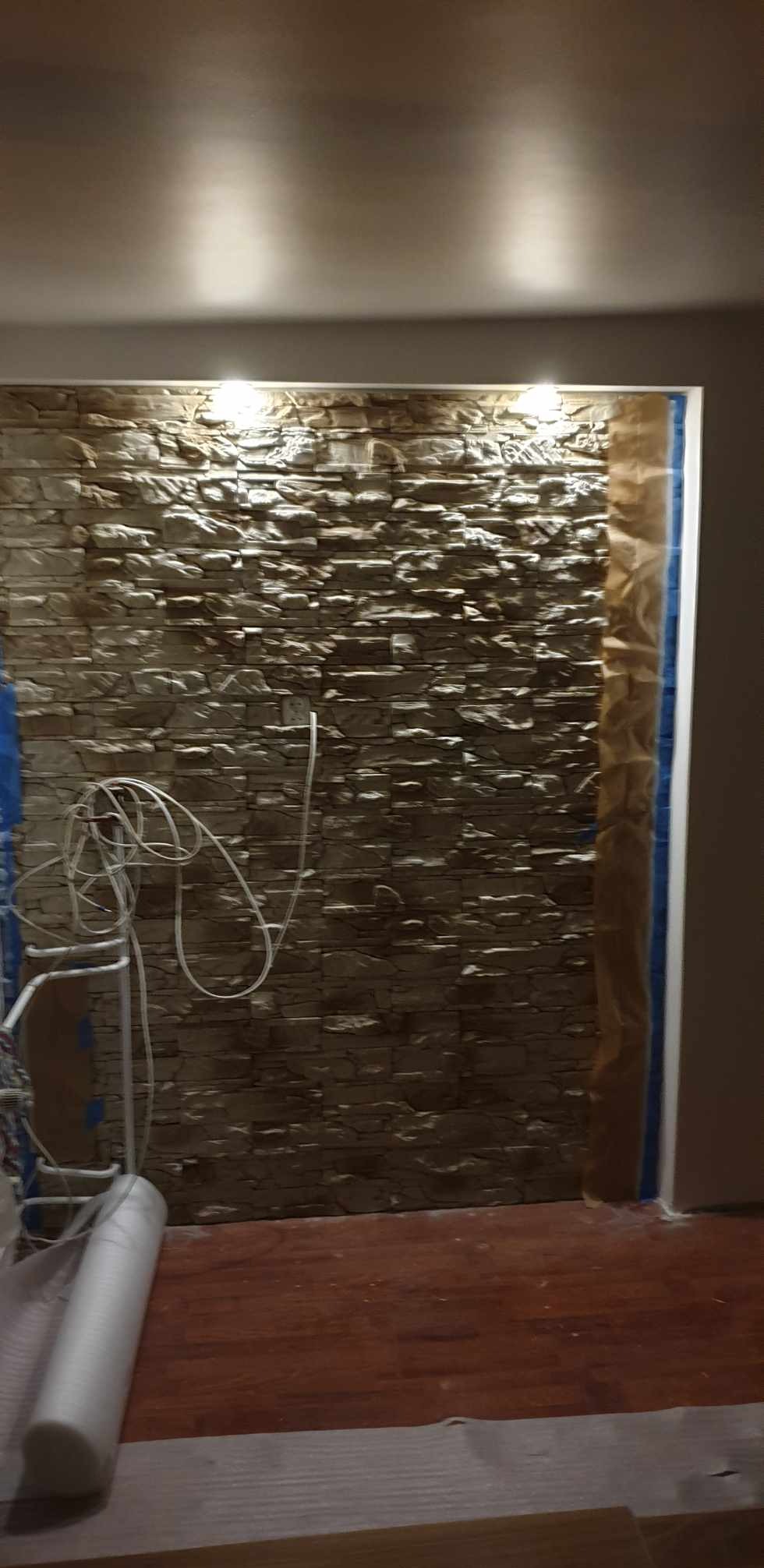Ściana z dekoracyjnego kamienia w niszy, zabezpieczona folią malarską, widoczne przewody elektryczne i elementy rusztowania.