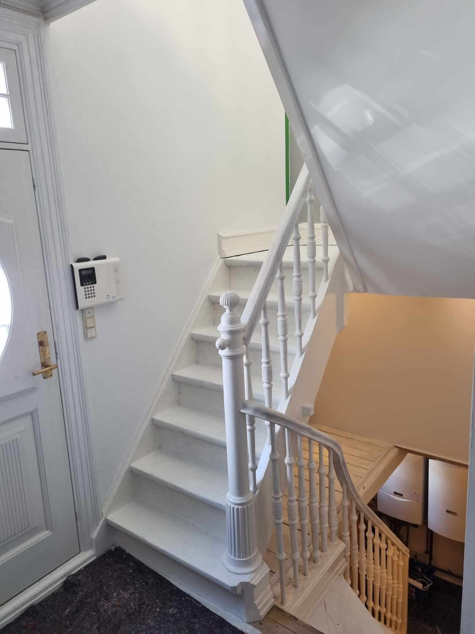 Białe schody z drewnianą balustradą w trakcie renowacji, widoczny fragment drzwi wejściowych i panel alarmowy.