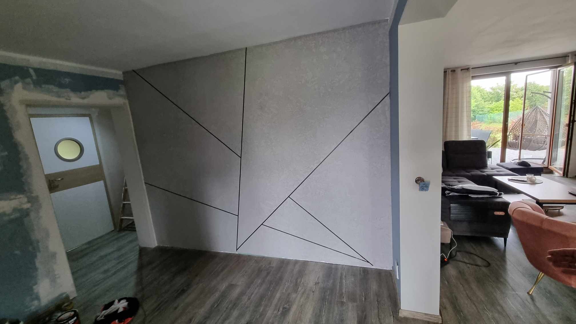 Ściana z gładzią w Chełmnie z geometrycznym wzorem z czarnych linii, widoczny fragment pokoju z drzwiami i salon z sofą w tle.