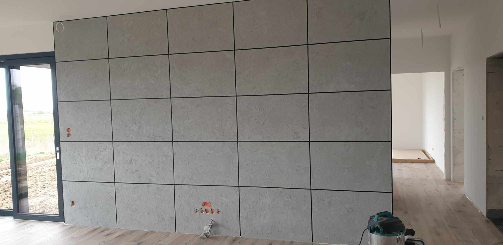 Ściana wykończona panelami dekoracyjnymi imitującymi beton z czarnymi fugami, widoczne gniazdka elektryczne przed montażem osprzętu, odkurzacz na podłodze, w tle widok na zewnątrz przez okno...