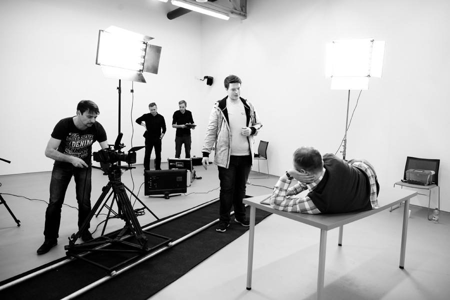 Czarno-białe zdjęcie przedstawia plan filmowy z operatorem przy profesjonalnej kamerze na szynach, oświetlaczami i aktorem leżącym na stole w trakcie nagrywania.