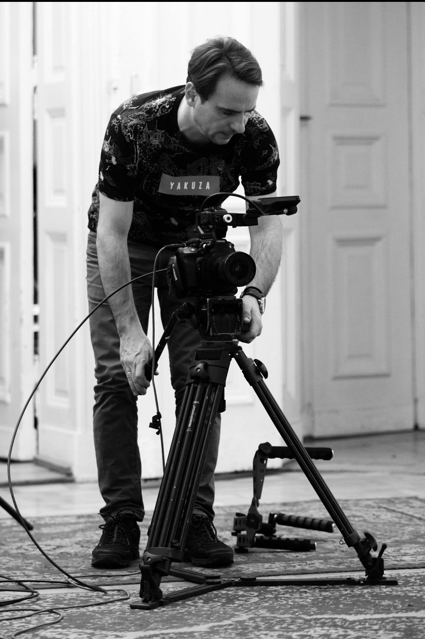 Czarno-białe zdjęcie filmowca w koszulce z napisem 'Yakuza', ustawiającego profesjonalną kamerę Canon na statywie w pomieszczeniu z białymi drzwiami w tle.