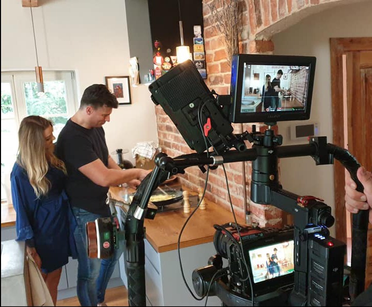 Ekipa filmowa kręci scenę w kuchni: mężczyzna przygotowuje posiłek na płycie grzewczej, kobieta stoi obok, widoczny profesjonalny sprzęt do filmowania z monitorem podglądowym.