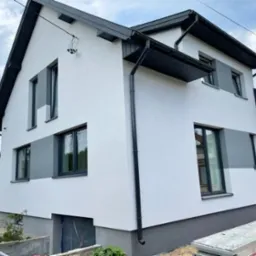 Dwukondygnacyjny dom po ociepleniu elewacji z szarą podmurówką i szarymi akcentami wokół okien, widoczne rynny i blaszane elementy dachu w kolorze grafitowym, częściowo odsłonięte wejście do piwnicy