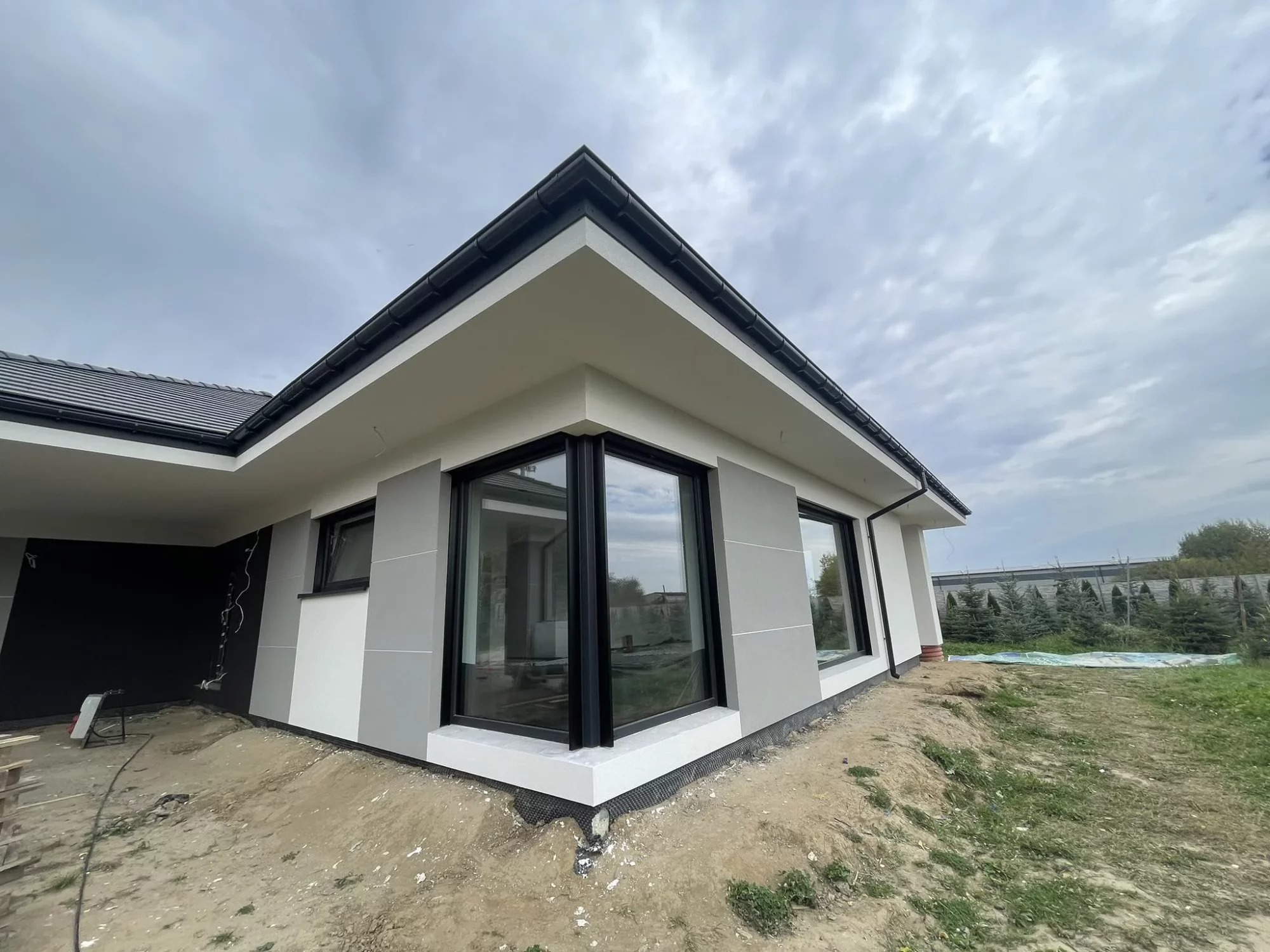 Nowoczesny dom jednorodzinny z szarą elewacją w trakcie wykańczania; widoczne duże okna z czarnymi ramami, fragment dachu pokrytego ciemną dachówką oraz niezagospodarowany teren wokół budynku.