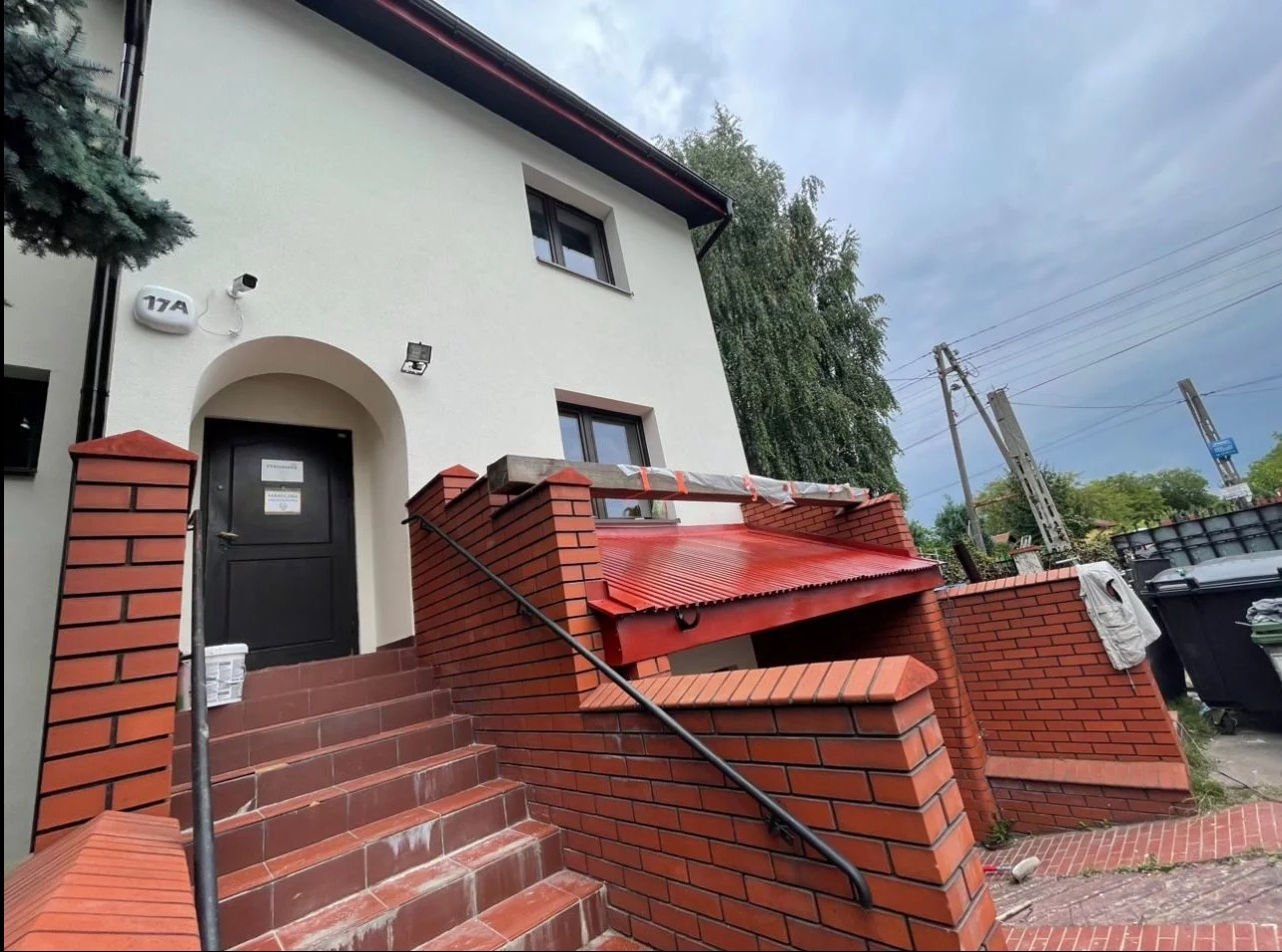 Dwupiętrowy budynek z białymi ścianami, ciemnymi oknami i wejściem z czerwonej cegły, nad którym widać zabezpieczoną konstrukcję, prawdopodobnie przed dalszymi pracami.