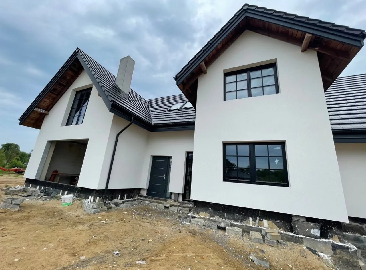 Nowoczesny dom jednorodzinny z białą elewacją i czarnymi ramami okiennymi, widok z dołu na ukończoną fasadę, teren wokół domu jeszcze w trakcie prac wykończeniowych, widoczne fundamenty...