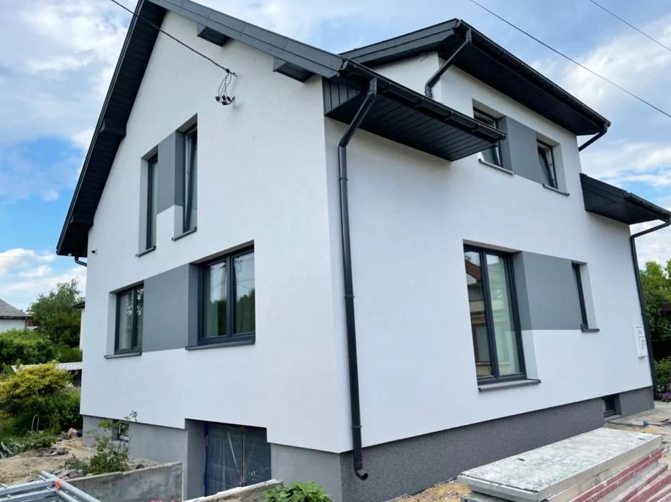 Dwukondygnacyjny dom po ociepleniu elewacji z szarą podmurówką i szarymi akcentami wokół okien, widoczne rynny i blaszane elementy dachu w kolorze grafitowym, częściowo odsłonięte wejście do piwnicy