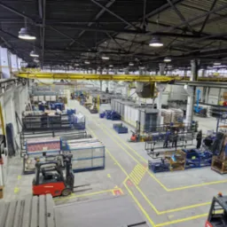 Szeroki kadr hali produkcyjnej w Wąchocku, z widocznymi wózkami widłowymi, różnymi elementami metalowymi przygotowanymi do obróbki oraz suwnicą pod stropem.