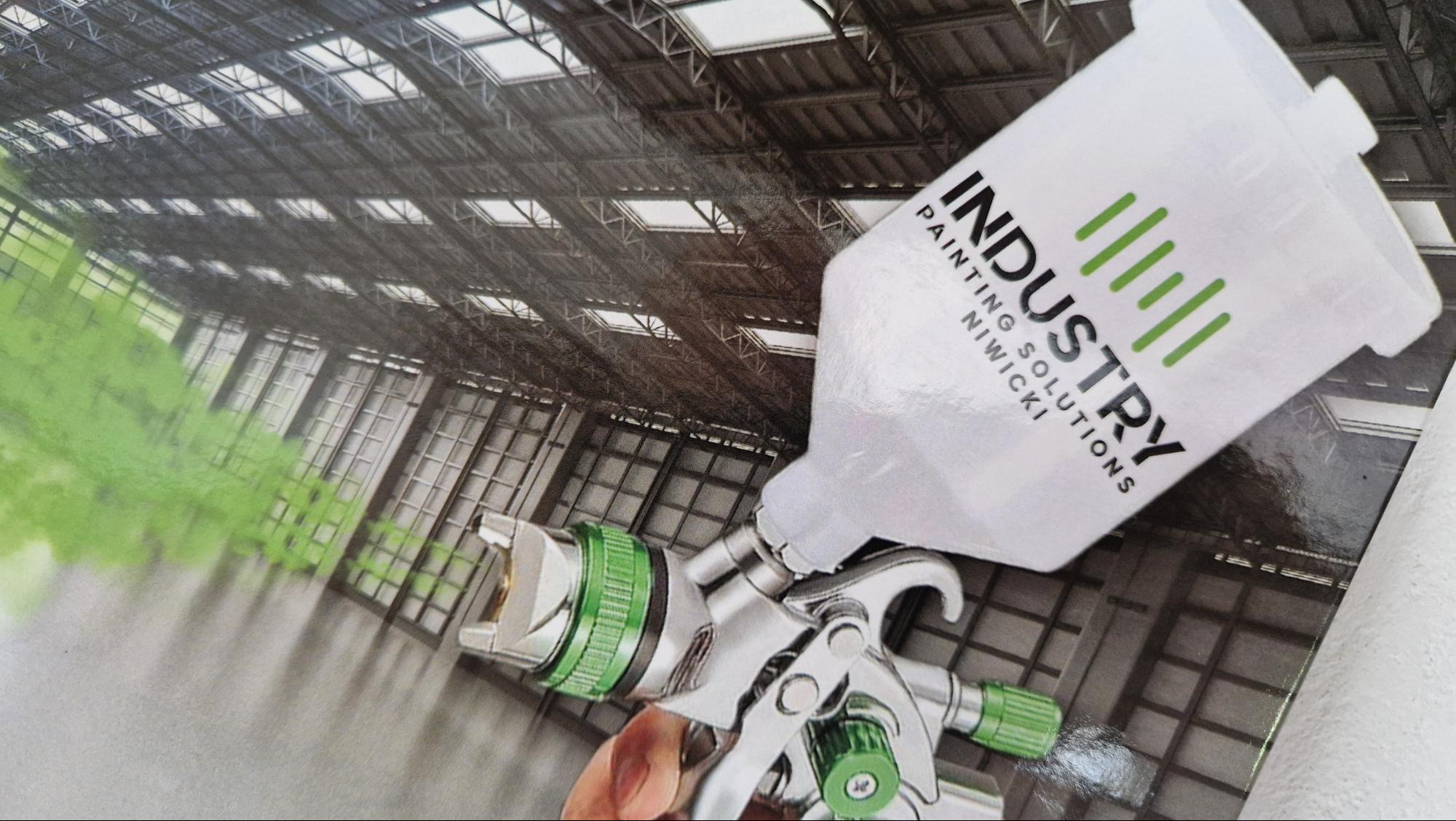 Pistolet do malowania proszkowego z logiem firmy Industry Painting Solutions Niwicki na tle zdjęcia hali przemysłowej.