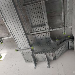 El-mont Volodymyr Bangula - Metalowe korytka kablowe z perforacją, zamontowane pod betonowym sufitem z elementami mocującymi w kolorze zielonym, tworzące system prowadzenia kabli.