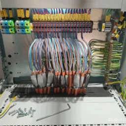 Rozdzielnica elektryczna w trakcie montażu: widoczne kable z oznaczeniami, listwy zaciskowe, bezpieczniki oraz narzędzia, w tym klucz imbusowy i śruby.