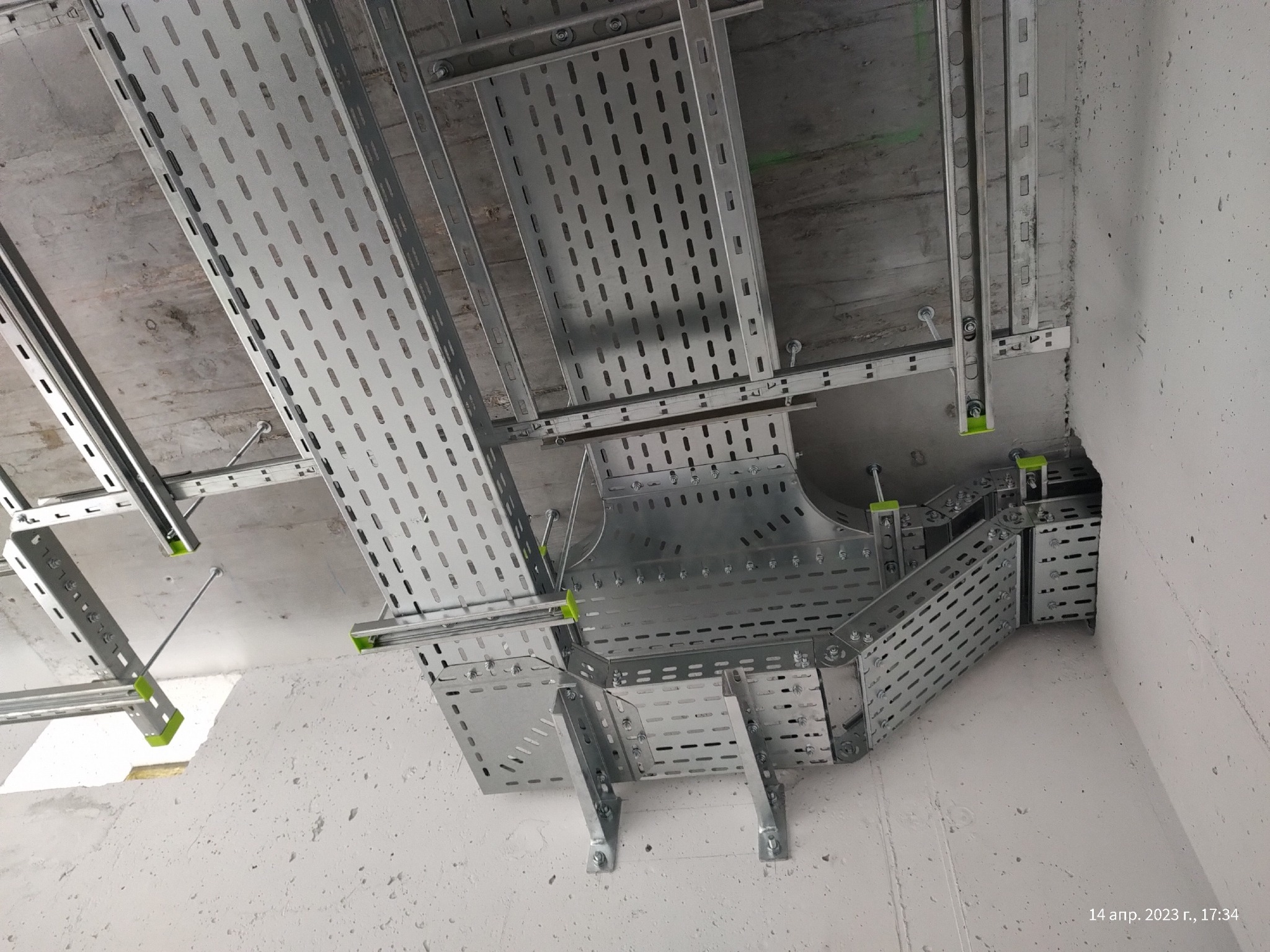 Metalowe korytka kablowe z perforacją, zamontowane pod betonowym sufitem z elementami mocującymi w kolorze zielonym, tworzące system prowadzenia kabli.