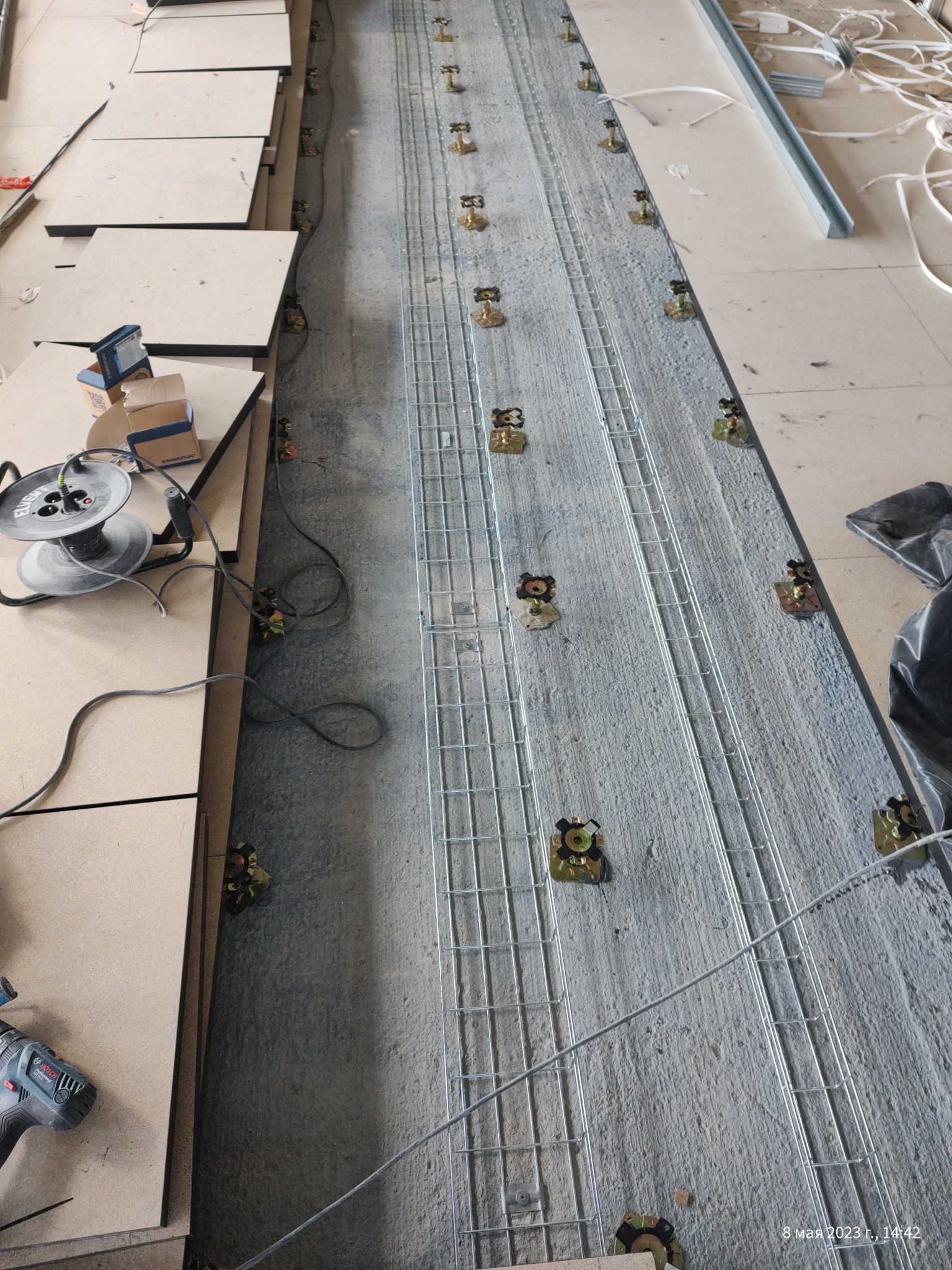 Montaż podłogi podniesionej: widok z góry na betonową posadzkę z zamocowanymi metalowymi wspornikami i siatką, obok leżą płyty podłogowe oraz narzędzia - wiertarka i zwijarka do kabli.