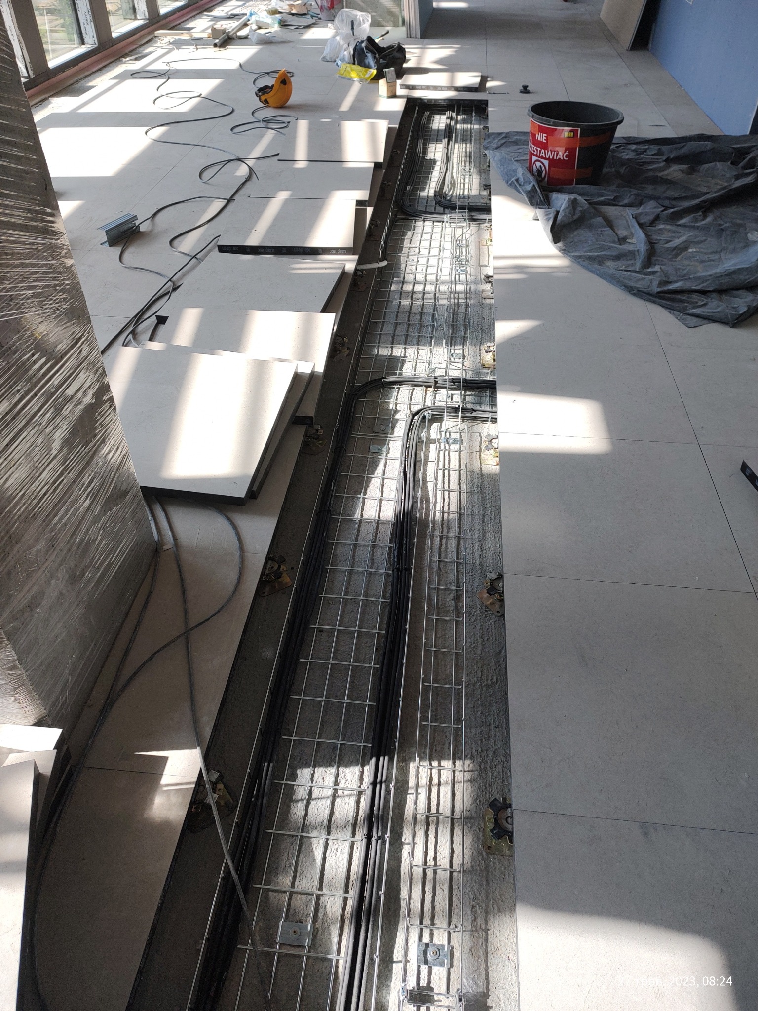 Wykonywanie podpodłogowej instalacji kablowej w nowym budynku w Warszawie. Widoczne koryto kablowe z ułożonymi przewodami, częściowo przykryte płytami podłogowymi, narzędzia pracy i wiadro...