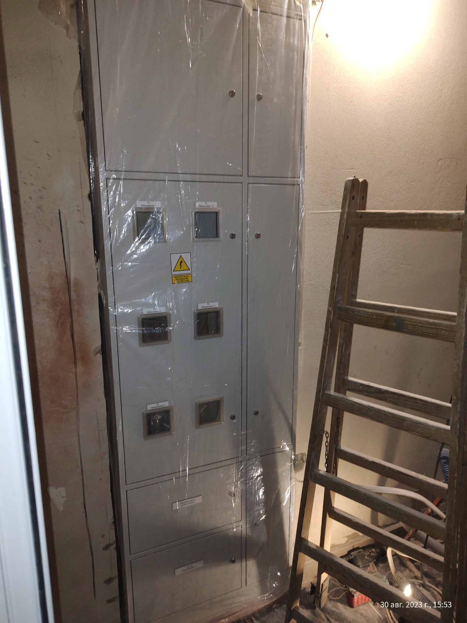 Nowa, szara rozdzielnica elektryczna, jeszcze zabezpieczona folią ochronną, umieszczona w surowym pomieszczeniu obok drewnianej drabiny, widoczne oznaczenia na panelach i ostrzeżenie o napięciu.