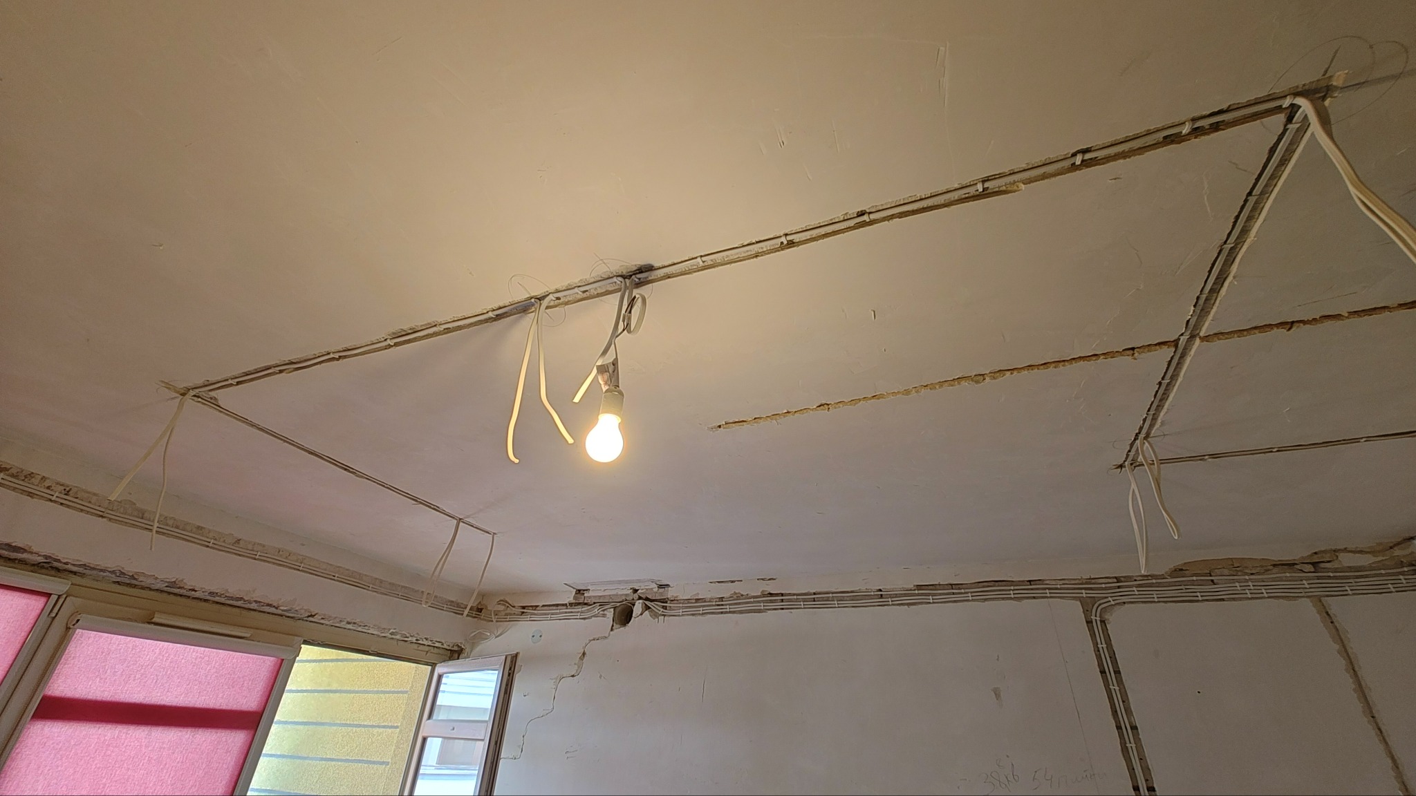 Widok sufitu w trakcie prac elektrycznych: kable w listwach przypodłogowych, tymczasowa żarówka na przewodach, widoczne ślady po kuciu w ścianach.