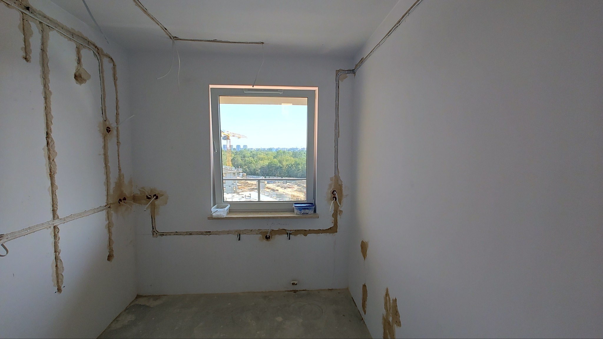 Wnętrze pokoju w trakcie prac elektrycznych: widoczne kable i puszki instalacyjne w bruzdach na ścianach, okno z widokiem na plac budowy.