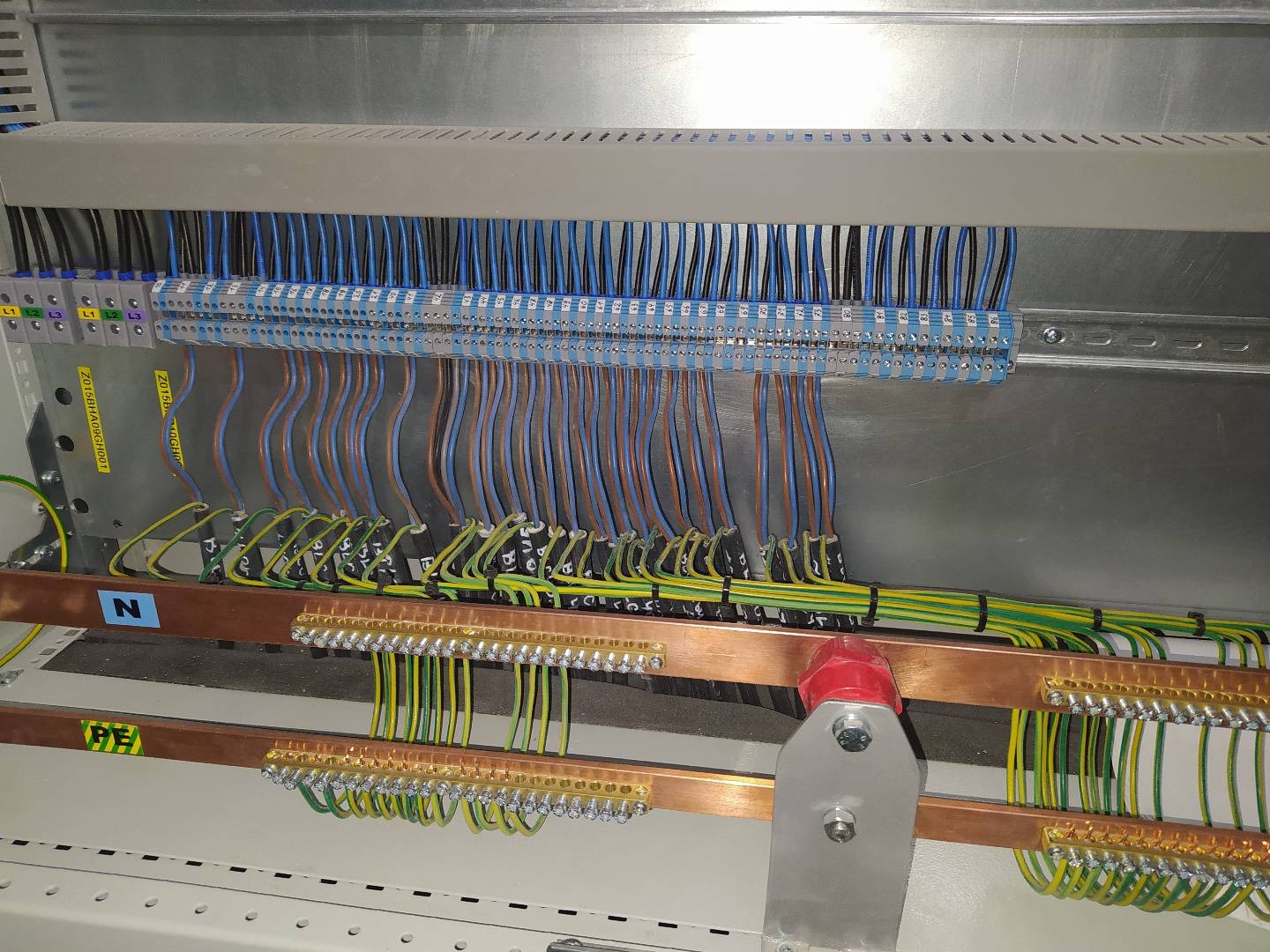 Wnętrze rozdzielni elektrycznej z uporządkowanymi przewodami o różnych kolorach (niebieskie, brązowe, żółto-zielone) podłączonymi do listew zaciskowych i szyn wyrównawczych, widoczne oznaczenia L1,...