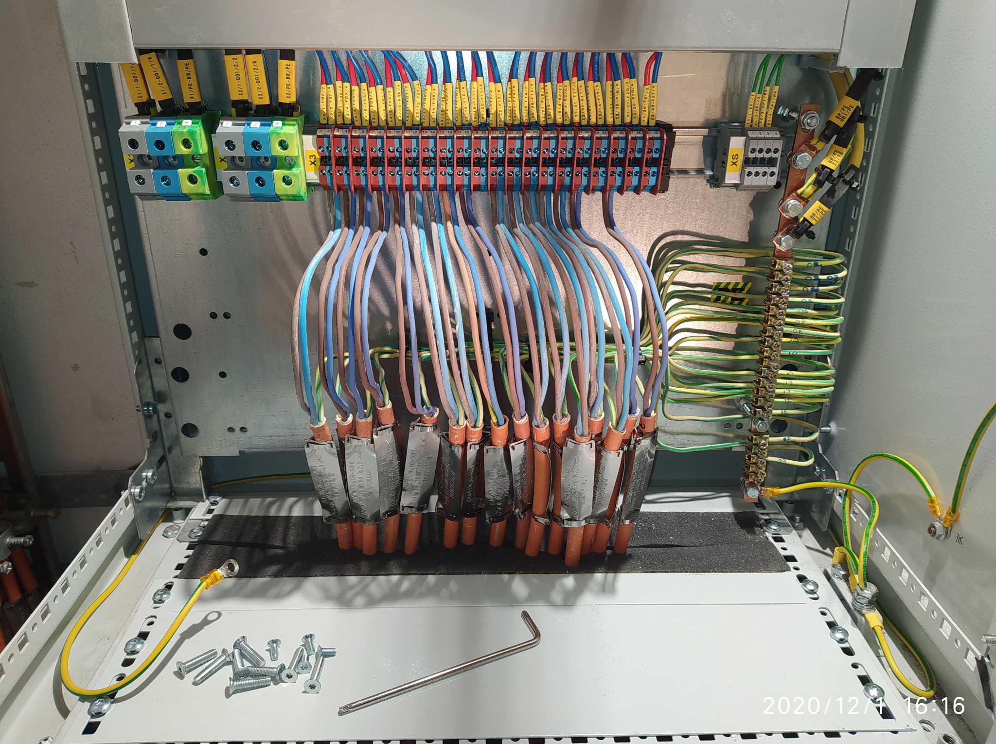 Rozdzielnica elektryczna w trakcie montażu: widoczne kable z oznaczeniami, listwy zaciskowe, bezpieczniki oraz narzędzia, w tym klucz imbusowy i śruby.