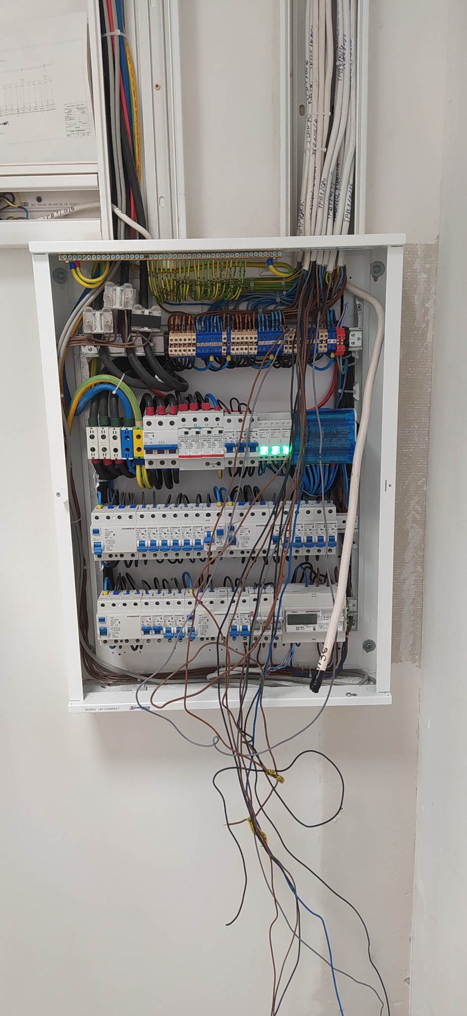 Otwarta skrzynka rozdzielcza z widocznymi bezpiecznikami, przewodami i licznikiem energii elektrycznej, niektóre przewody zwisają poza skrzynkę, widoczne zielone diody na zabezpieczeniach...