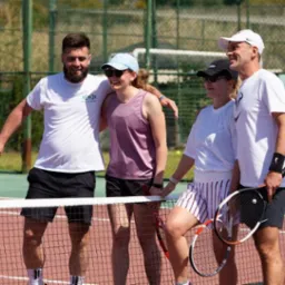 Grupa czterech uśmiechniętych osób, w tym dwie kobiety, stoi na korcie tenisowym trzymając rakiety po zakończonej grze, widok zza siatki.