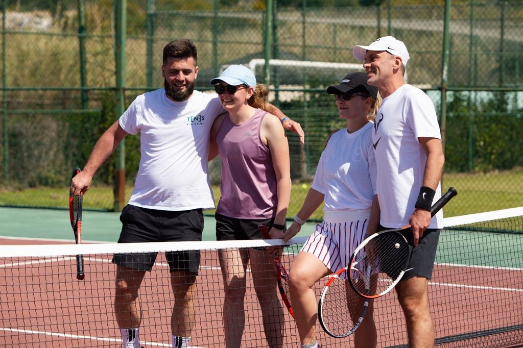 Grupa czterech uśmiechniętych osób, w tym dwie kobiety, stoi na korcie tenisowym trzymając rakiety po zakończonej grze, widok zza siatki.