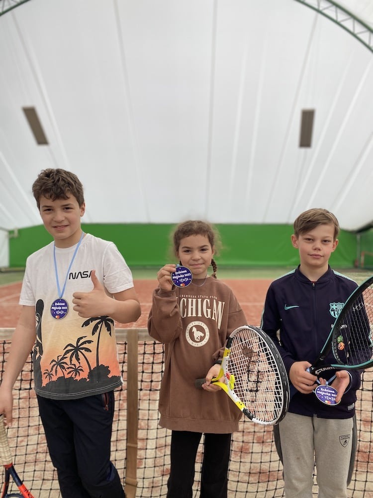 Trójka dzieci w Jawczycach z rakietami tenisowymi i medalami po turnieju tenisowym, na tle kortu pod zadaszeniem.