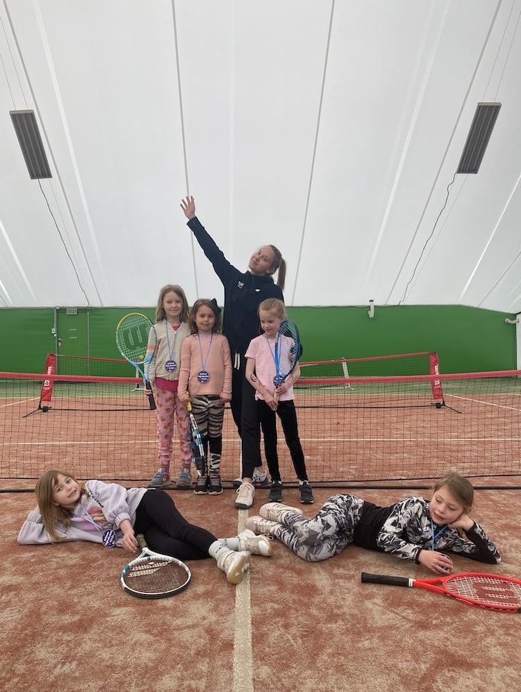 Grupa dzieci z trenerką pozuje z rakietami tenisowymi na korcie w hali, część dzieci leży na korcie, a część stoi za siatką, wszystkie dzieci mają przypięte identyfikatory.
