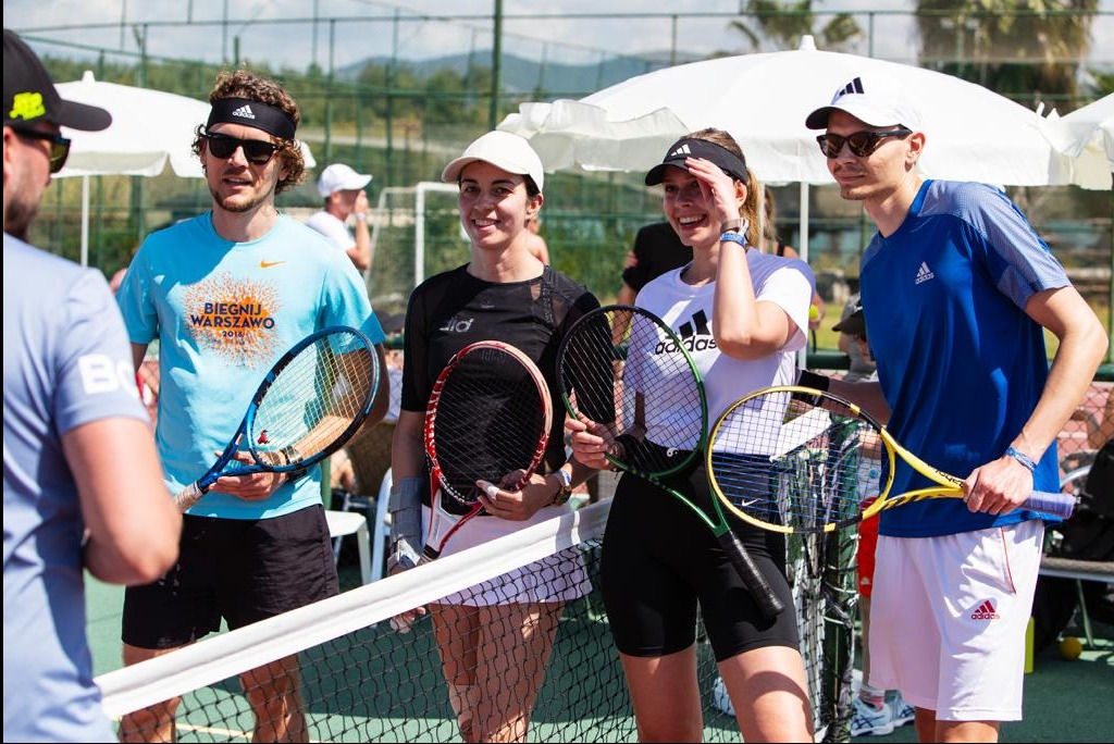 Grupa młodych ludzi w strojach sportowych, z rakietami tenisowymi, stoi przy siatce tenisowej na tle kortu i białych parasoli przeciwsłonecznych.