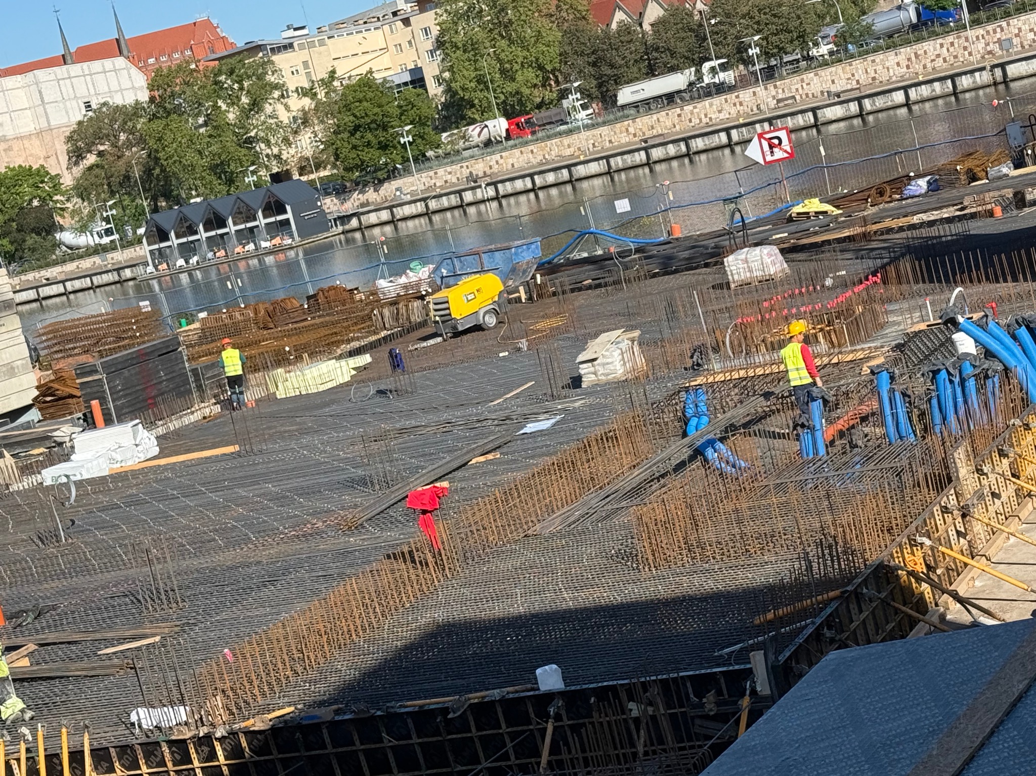 Rozległy teren budowy z widoczną siatką zbrojeniową, pracownikami w żółtych kamizelkach i sprzętem budowlanym na tle rzeki i miejskiej zabudowy.