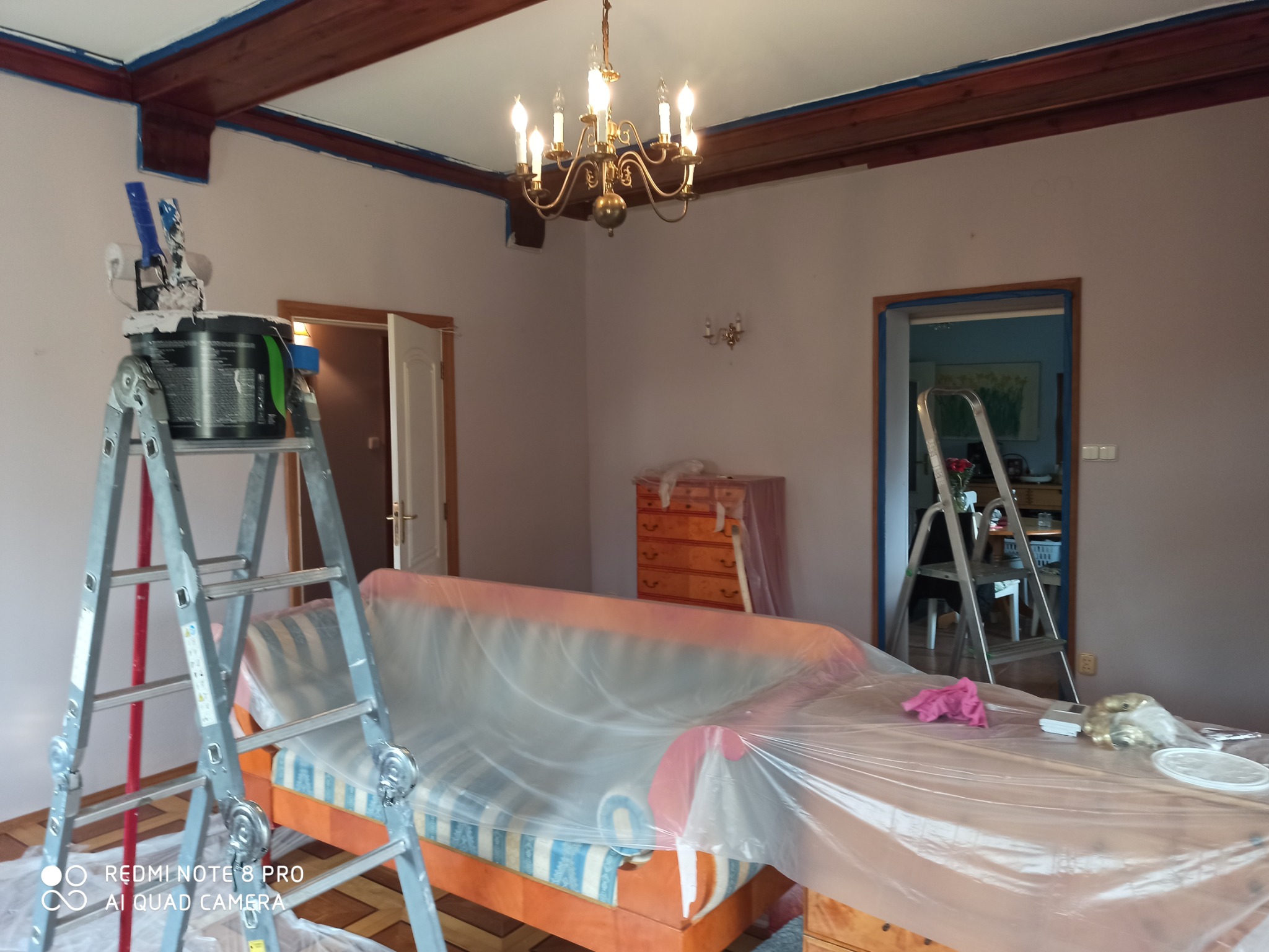 Pokój w trakcie odświeżania: ściany pomalowane na jasny kolor, drewniany sufit z widocznymi belkami zabezpieczony taśmą malarską, meble zakryte folią ochronną, drabina z wiadrem farby i wałkami...