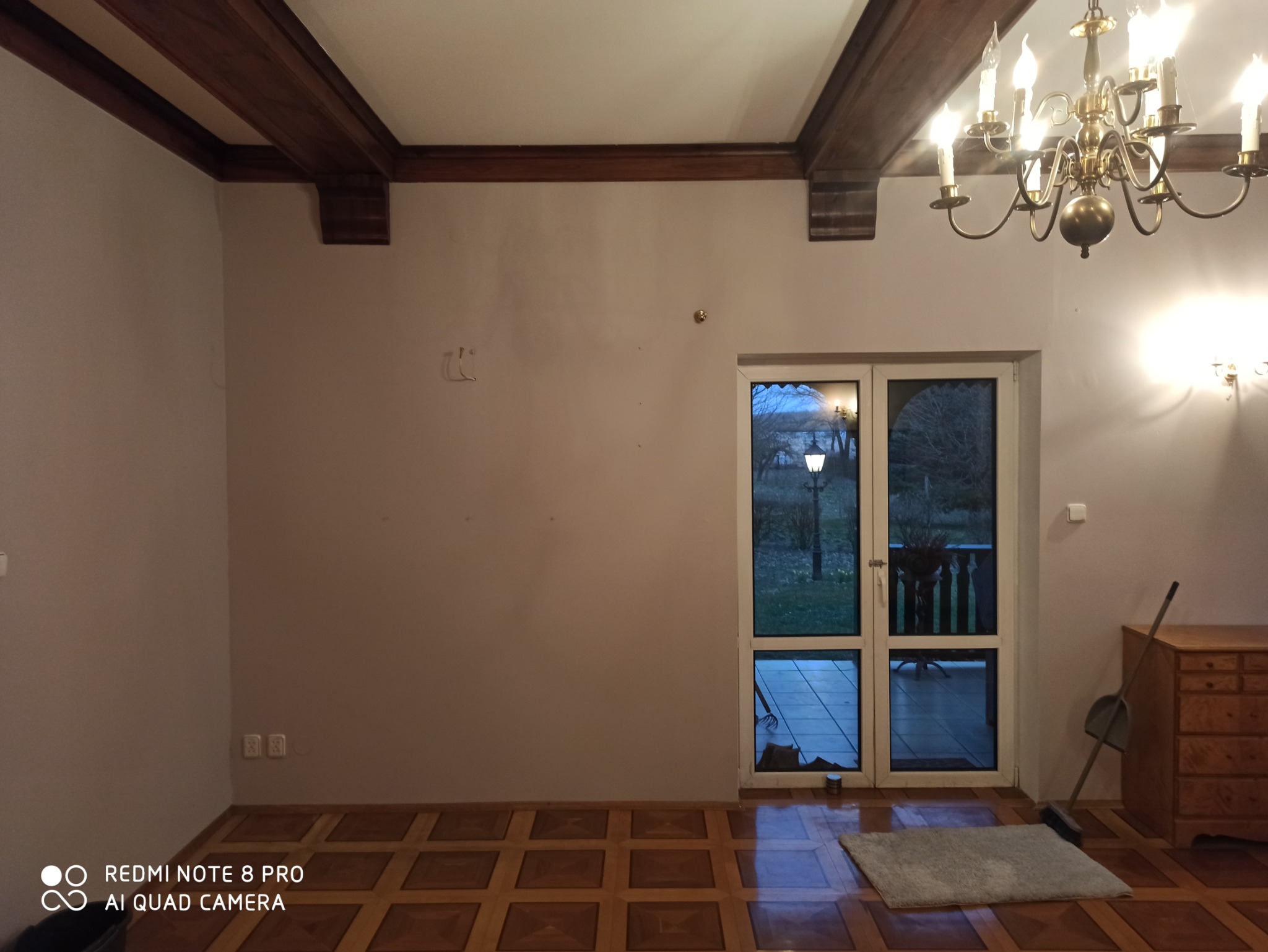 Pokój przed remontem z drewnianymi belkami sufitowymi, parkietem i widokiem na oświetlony lampion na zewnątrz przez przeszklone drzwi balkonowe.