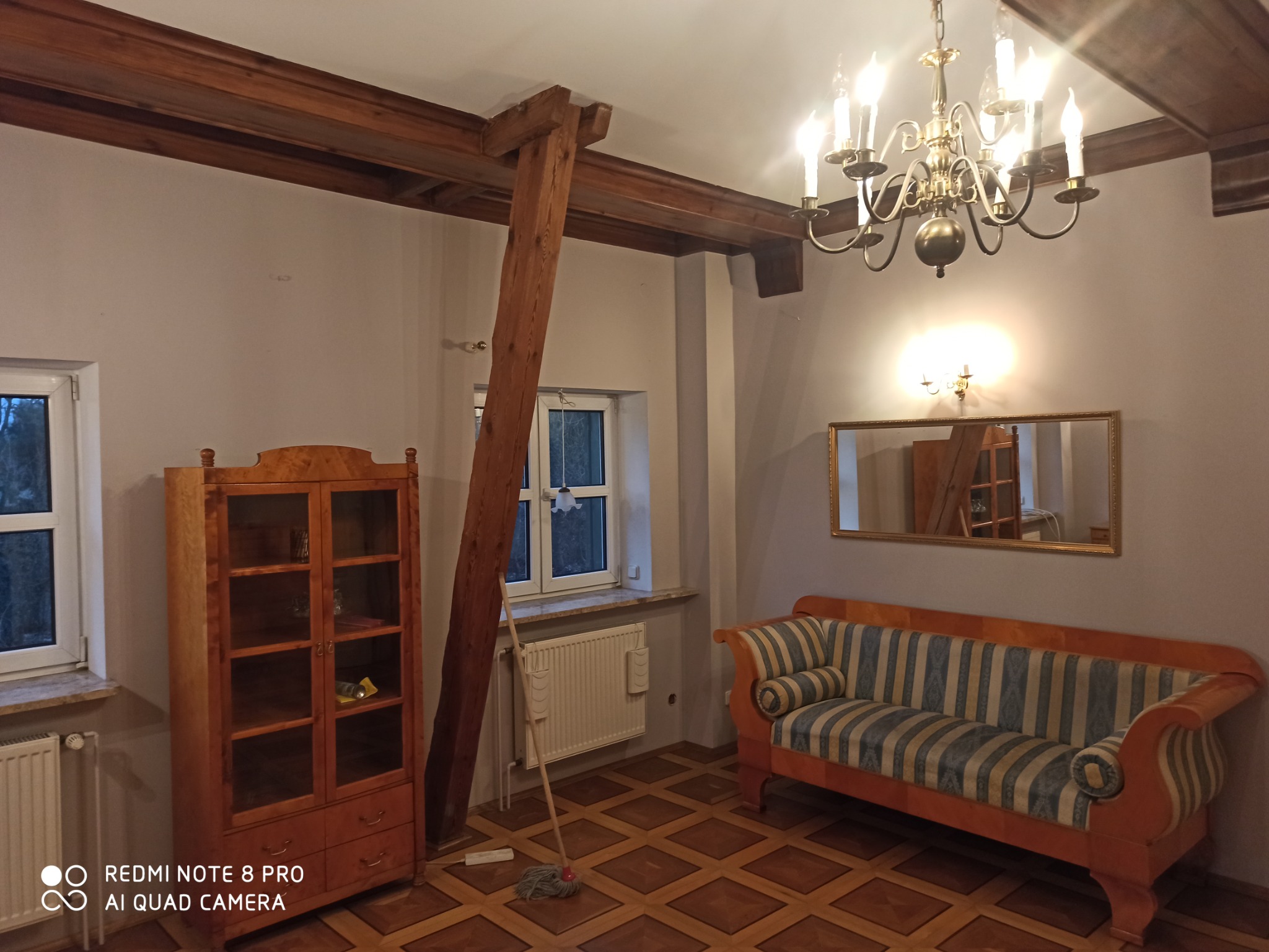Pokój z drewnianą podłogą w kwadraty, drewnianą witryną, kanapą w paski, lustrem w złotej ramie i drewnianą belką podpierającą sufit, widoczne ślady po malowaniu na ścianie.