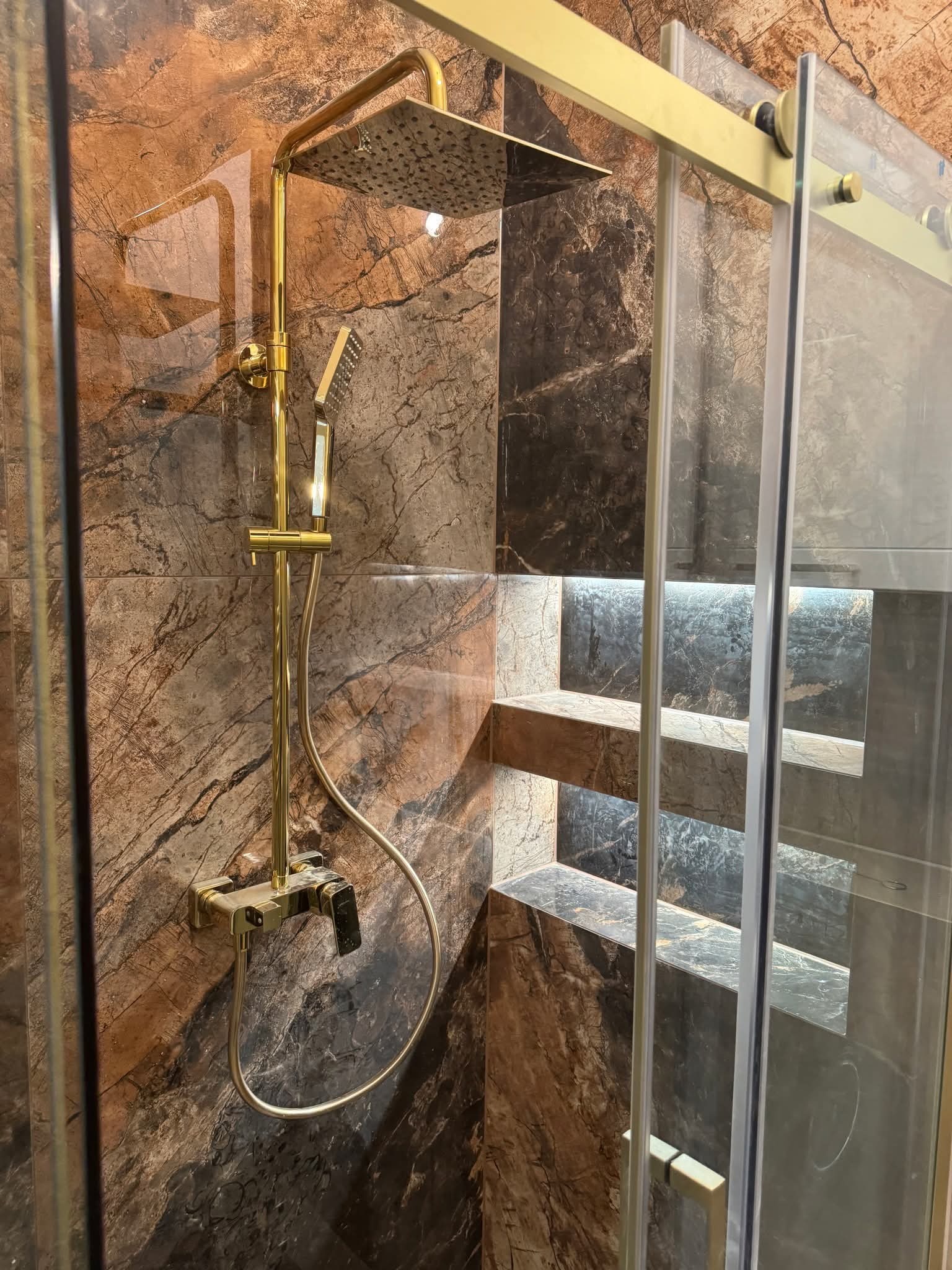 Nowoczesna kabina prysznicowa z deszczownicą w złotym kolorze, marmurowe płytki i półki z podświetleniem LED. Eleganckie wykończenie łazienki.