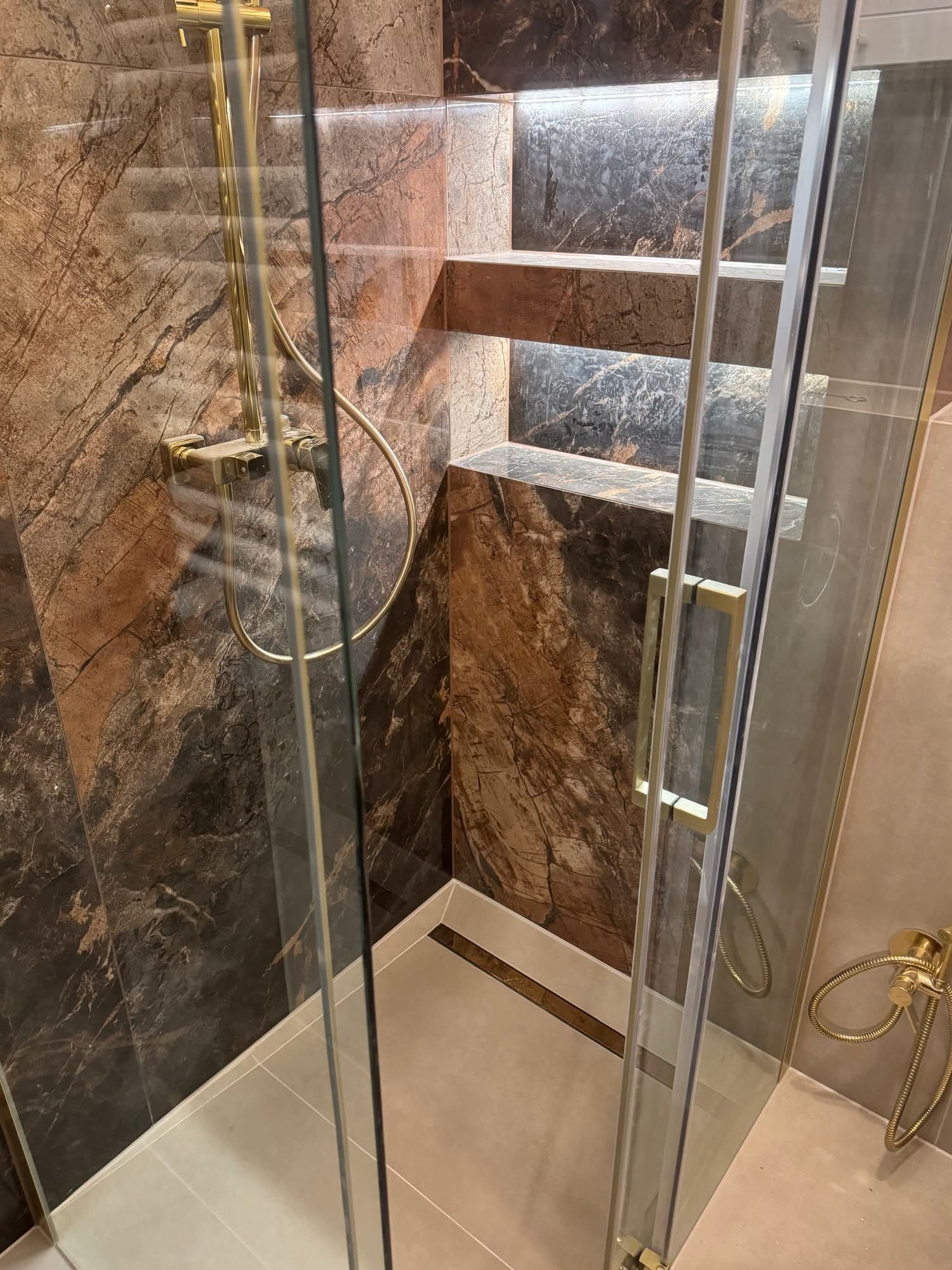 Luksusowa kabina prysznicowa z marmurowymi płytkami, złotą armaturą i podświetlanymi półkami. Nowoczesny design łazienki z dbałością o detale.