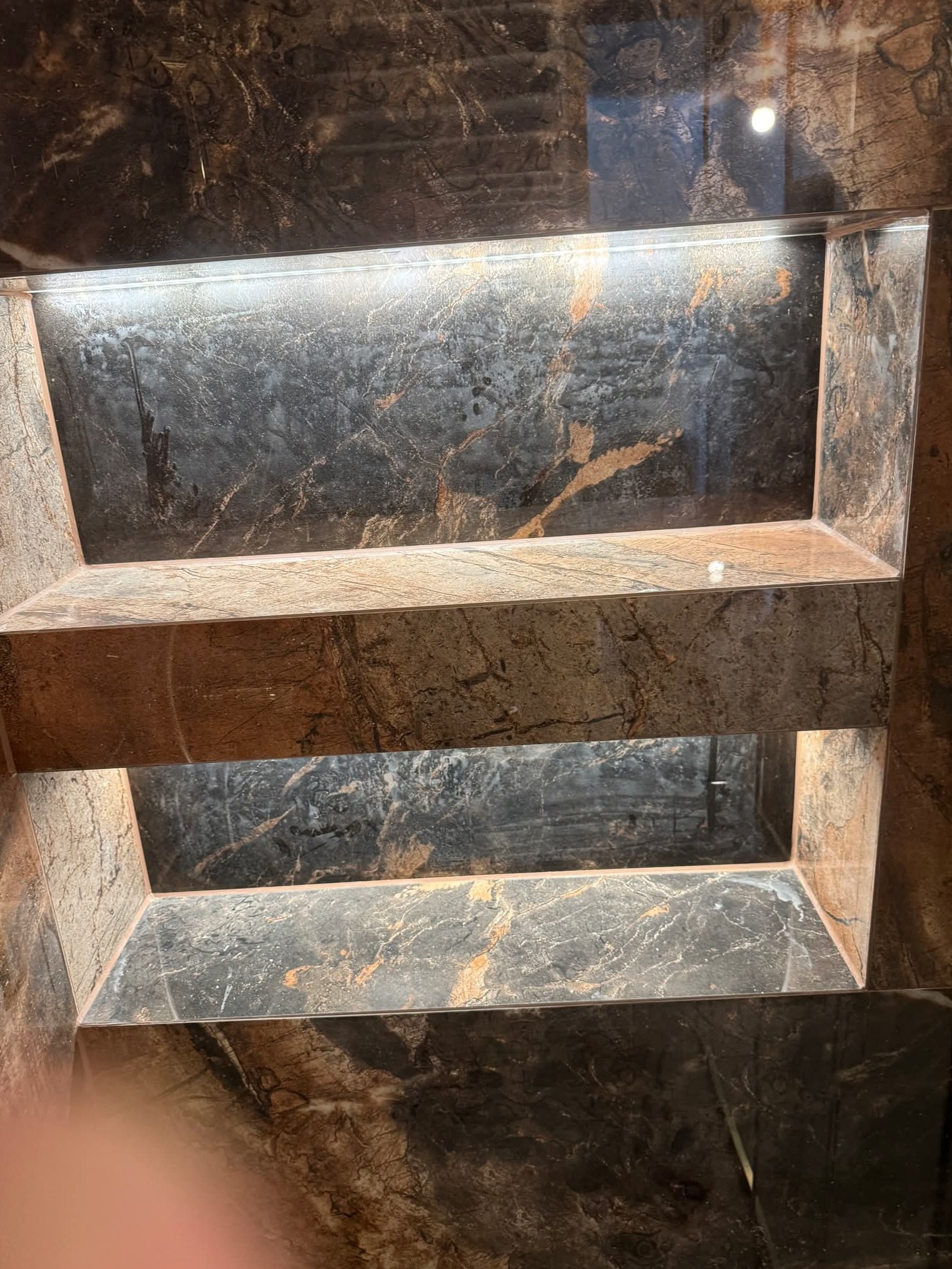 Wykończona wnęka w łazience z marmurowymi płytkami w odcieniach brązu i szarości, podświetlona listwą LED. Widok z dołu, ukazujący precyzję wykonania.