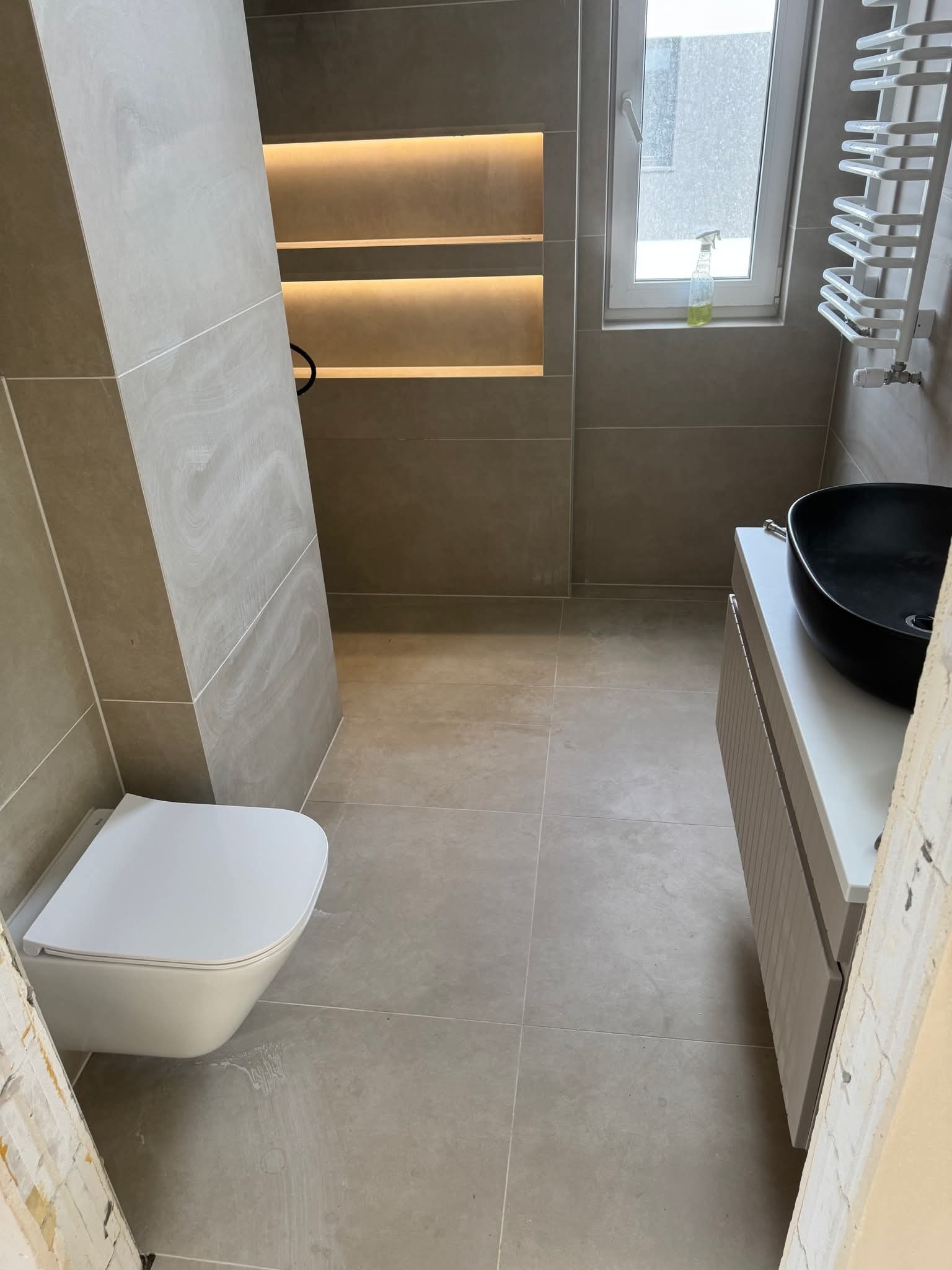 Wykończona łazienka z beżowymi płytkami, podwieszaną toaletą i czarną umywalką na szafce. W tle grzejnik drabinkowy i wnęka z oświetleniem LED.