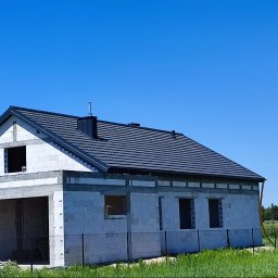 AWA-DACH Adam Pydyś - Nowy dom w trakcie budowy, z ciemnoszarą dachówką i kominem, widoczne otwory okienne i drzwiowe, ogrodzony siatką, z niebieskimi workami w garażu i dostawczym samochodem w tle.